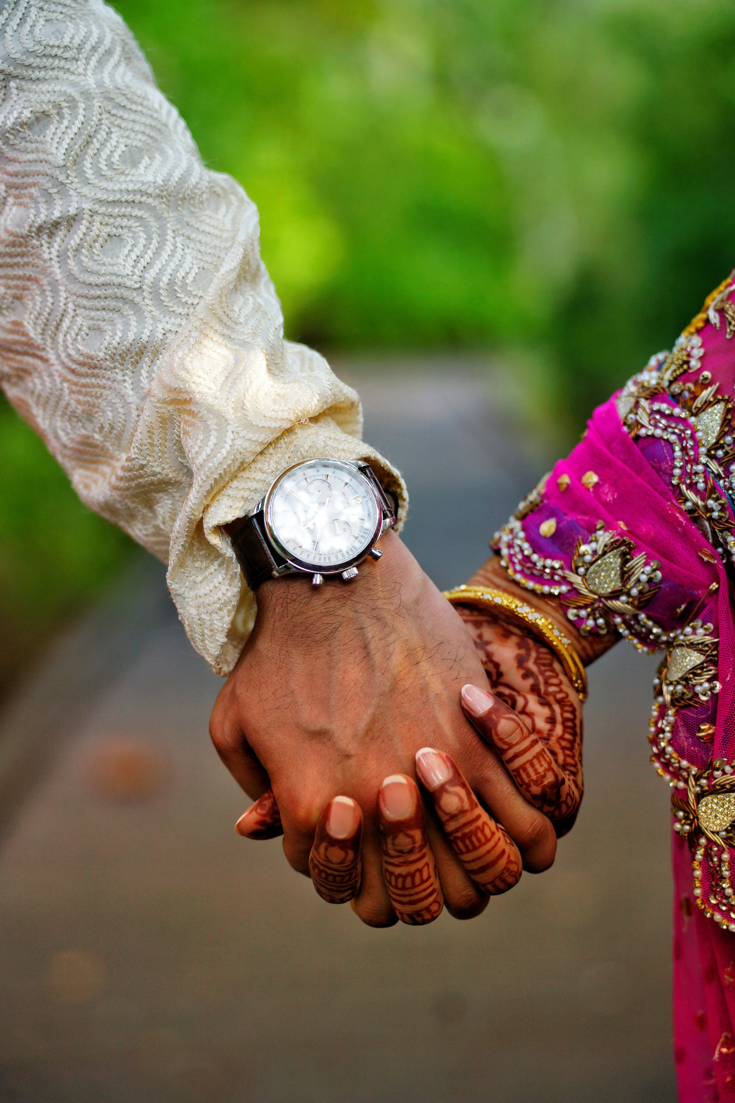 Close-up of two people holding hands, one wearing a traditional white embroidered garment with a watch, and the other wearing vibrant pink traditional attire decorated with gold and pearl embellishments. Both have intricate henna designs on their han