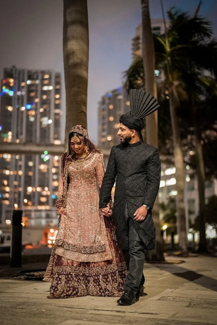 A couple dressed in traditional Indian wedding attire holding hands, walking outdoors at night in a city with palm trees and illuminated buildings in the background.