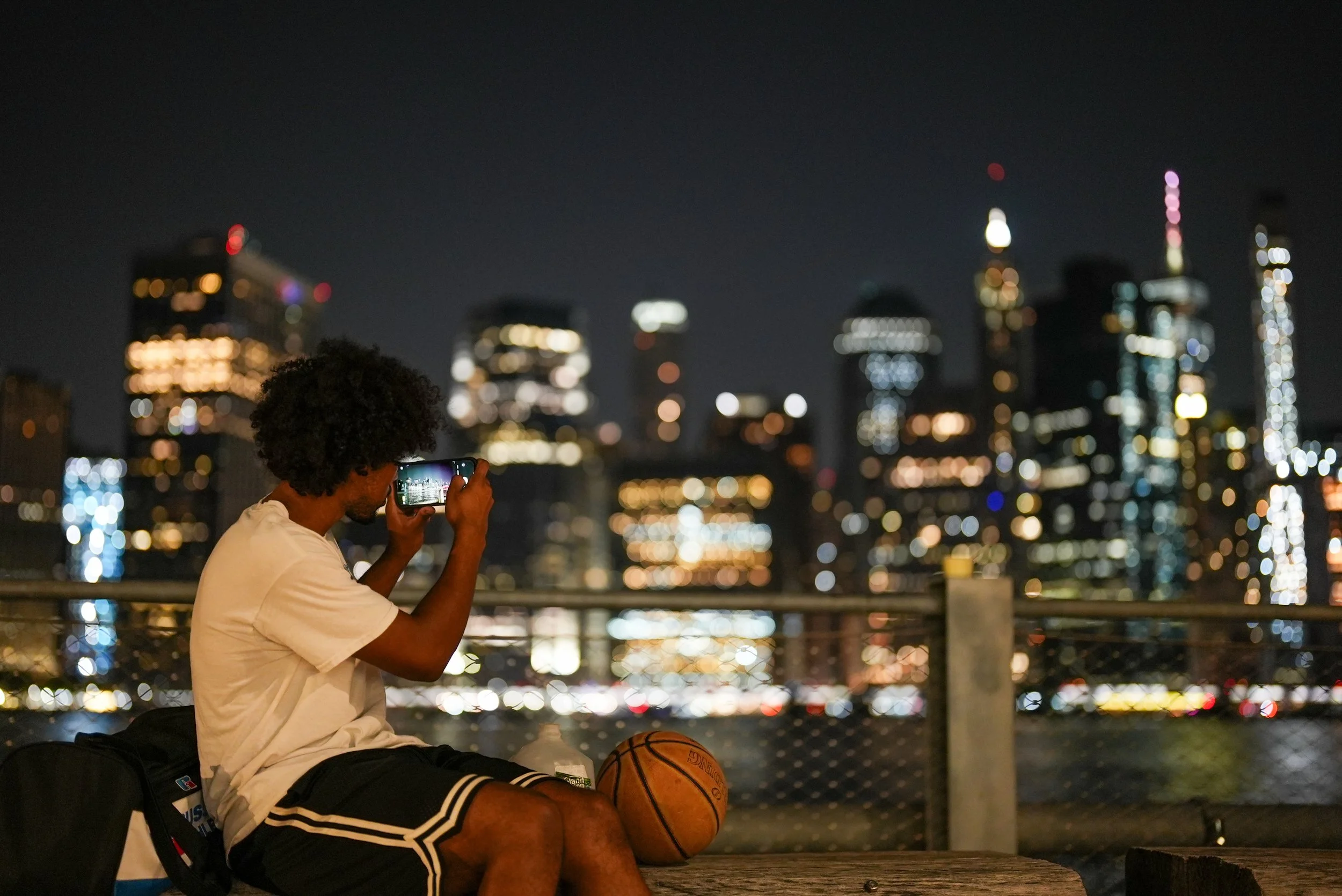 A man sitting on a bench at night, taking a photo of a city skyline with his phone. He has a basketball in his lap, and a backpack and basketball shoes are visible nearby.
