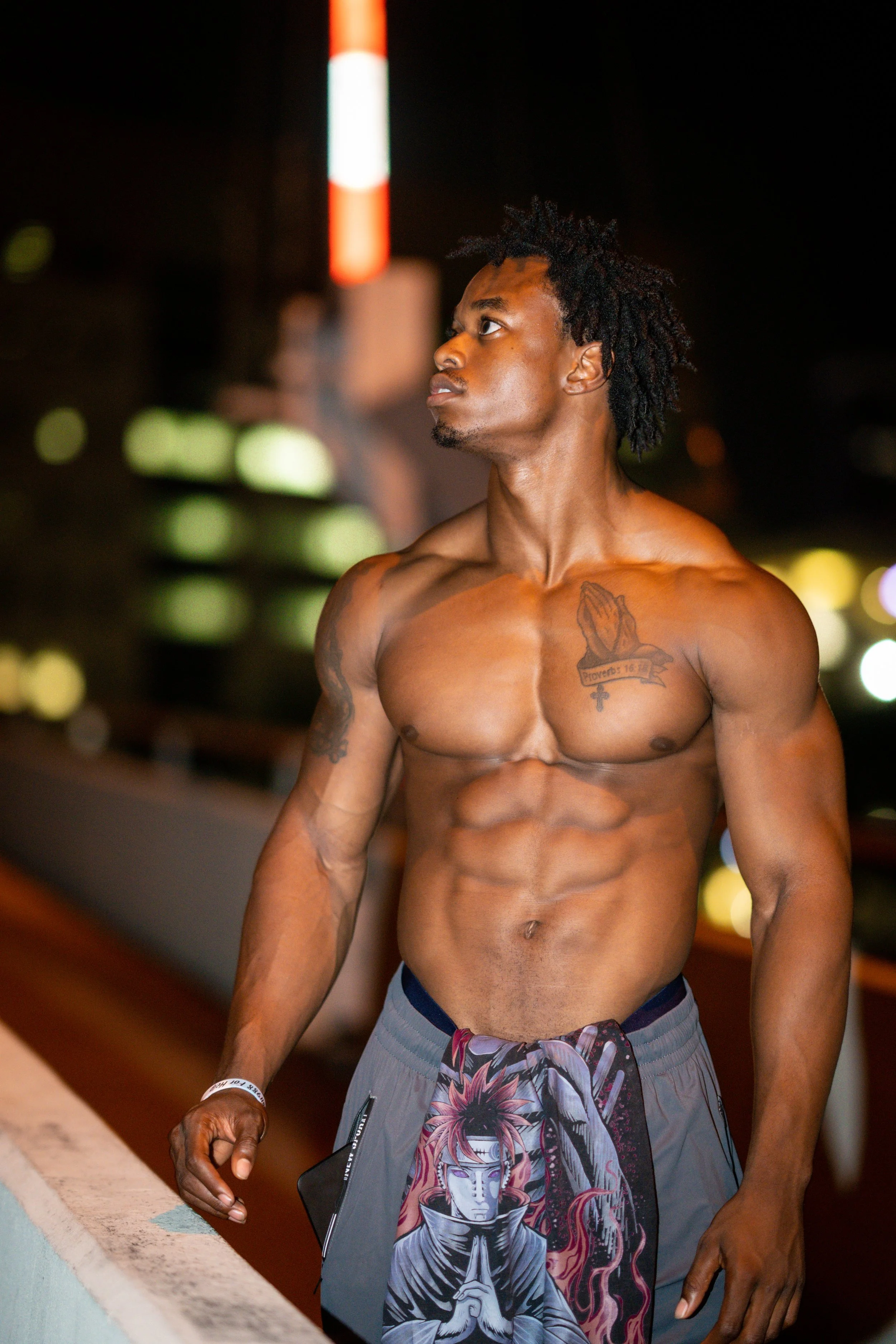 A shirtless man with tattoos on his chest and arm, wearing patterned shorts, standing at night near a low wall with city lights in the background.