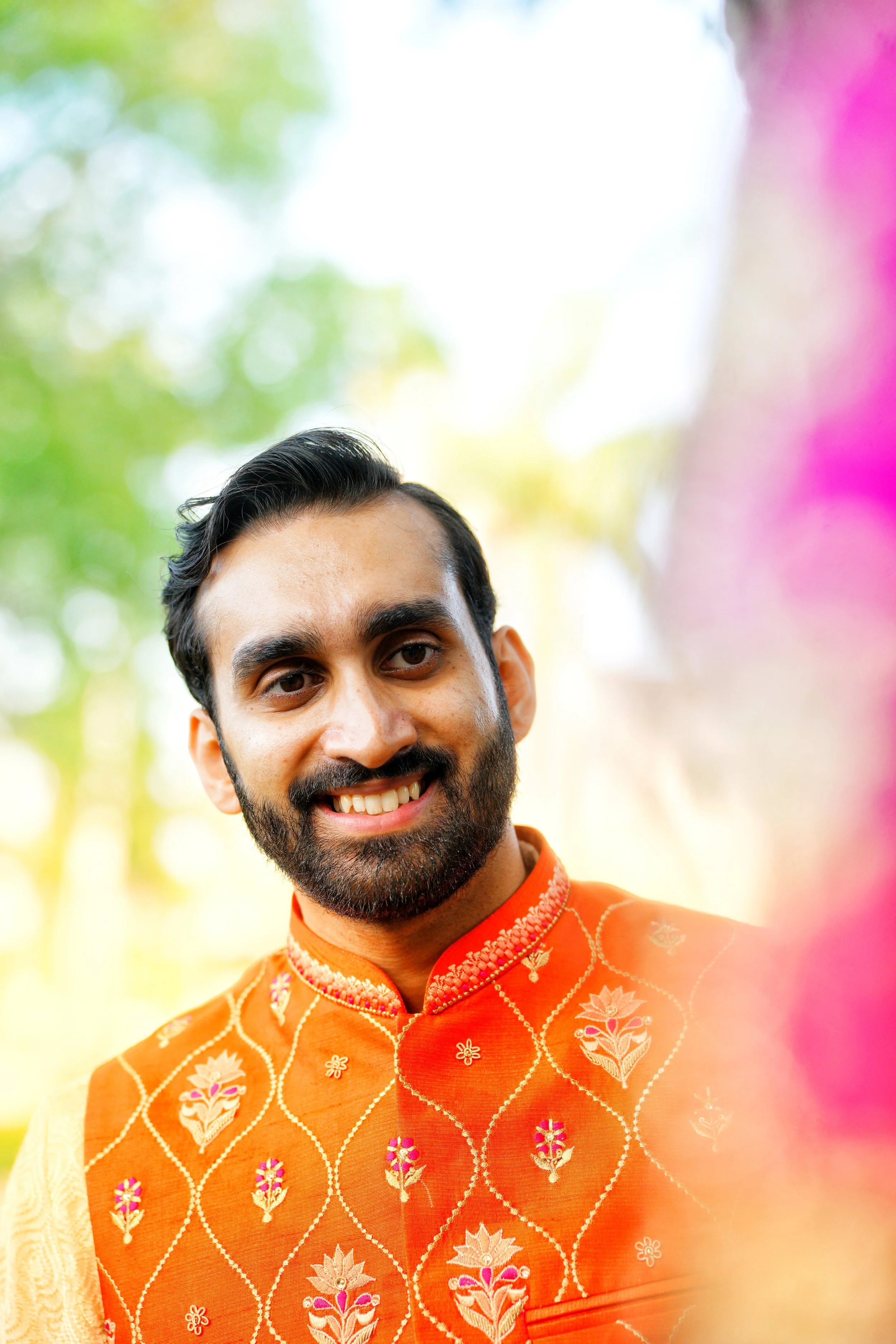 A man with dark hair and a beard smiling, wearing a bright orange traditional Indian kurta with intricate floral embroidery, outdoors with blurred colorful background.