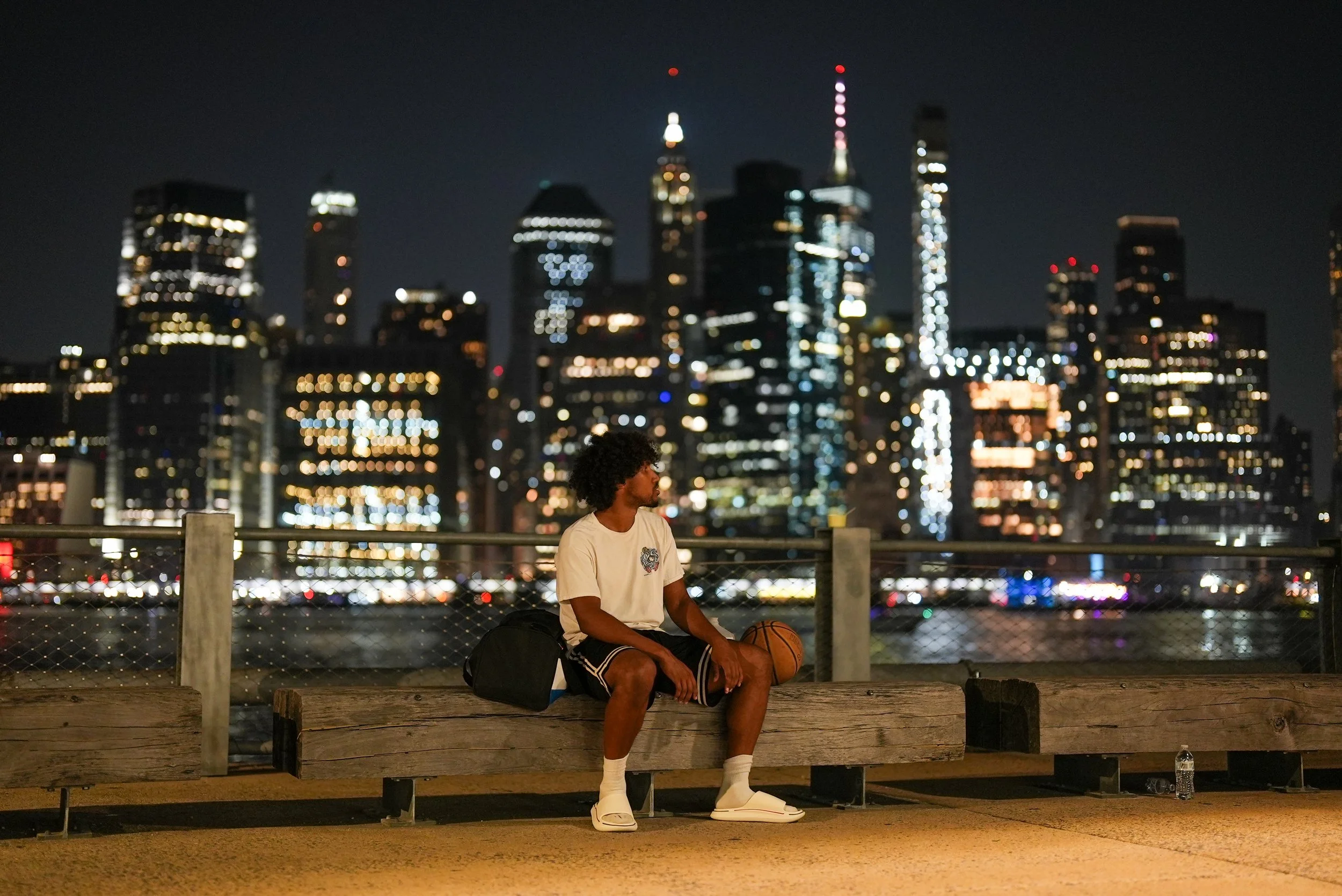 A young man sitting on a bench with a basketball and a backpack at night, city skyline with tall illuminated buildings in the background.