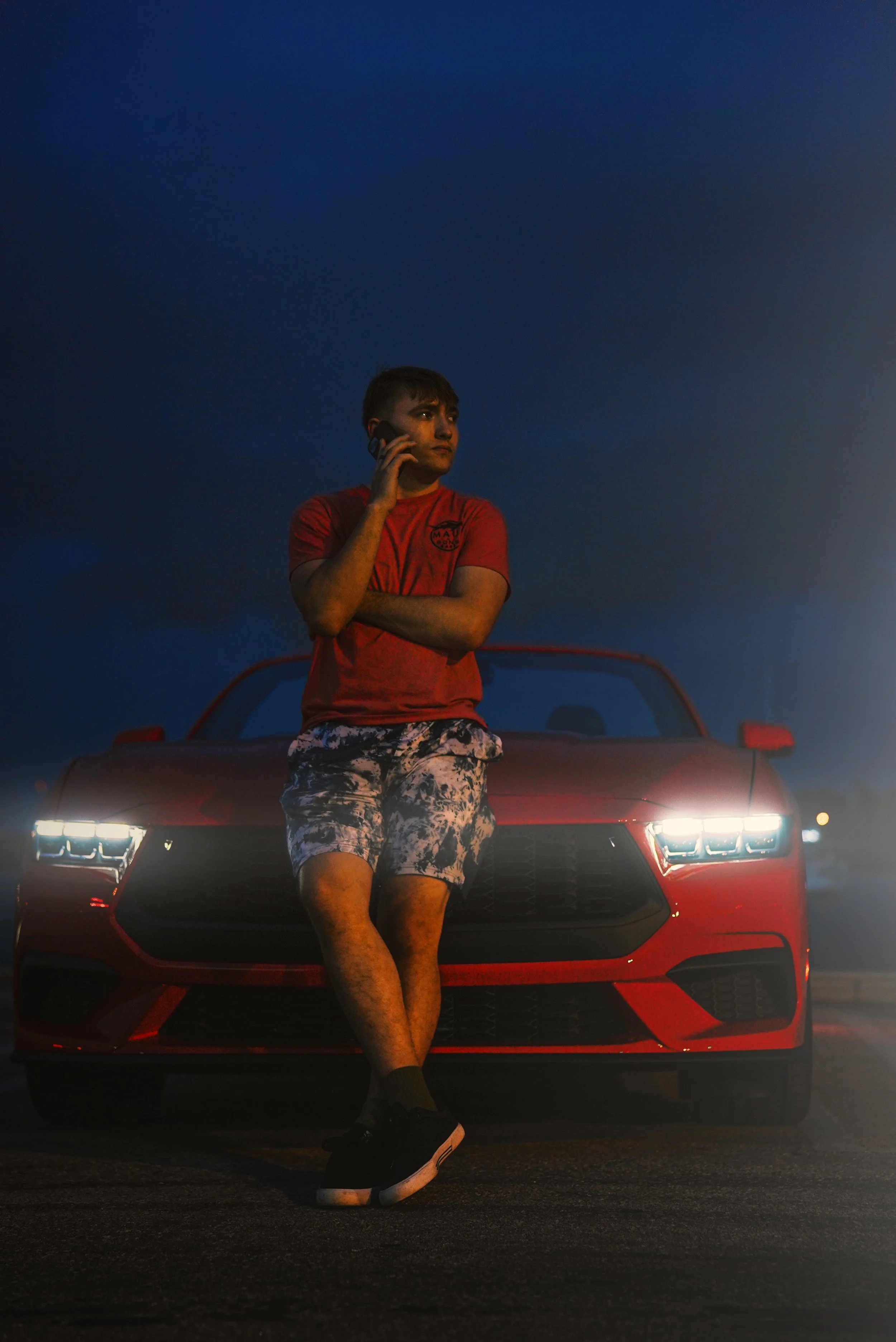 A young man standing in front of a red sports car, talking on his phone during twilight, with his arms crossed and wearing a red shirt and patterned shorts.