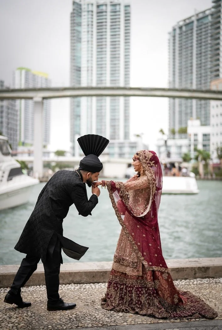 A man in traditional South Asian attire and a woman in an ornate wedding gown are by a waterway in an urban area with high-rise buildings. The man is kissing the woman's hand as she smiles at him.