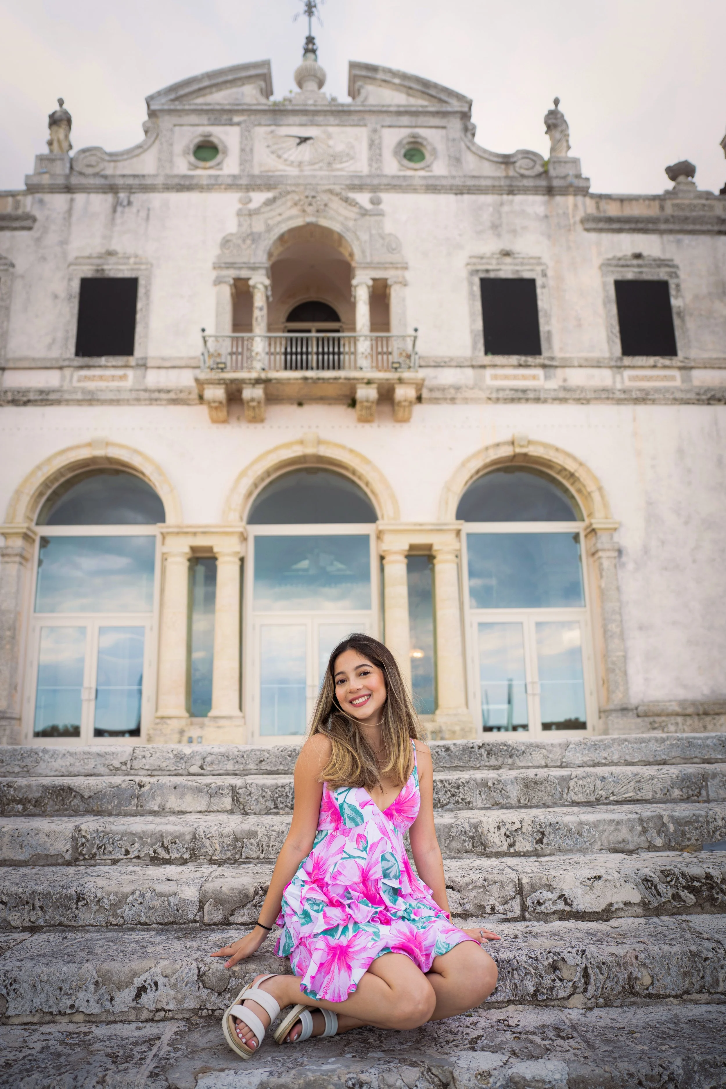 A young woman in a pink floral dress sitting on stone steps in front of an old stone building with large arched windows and a decorative upper facade, smiling at the camera.