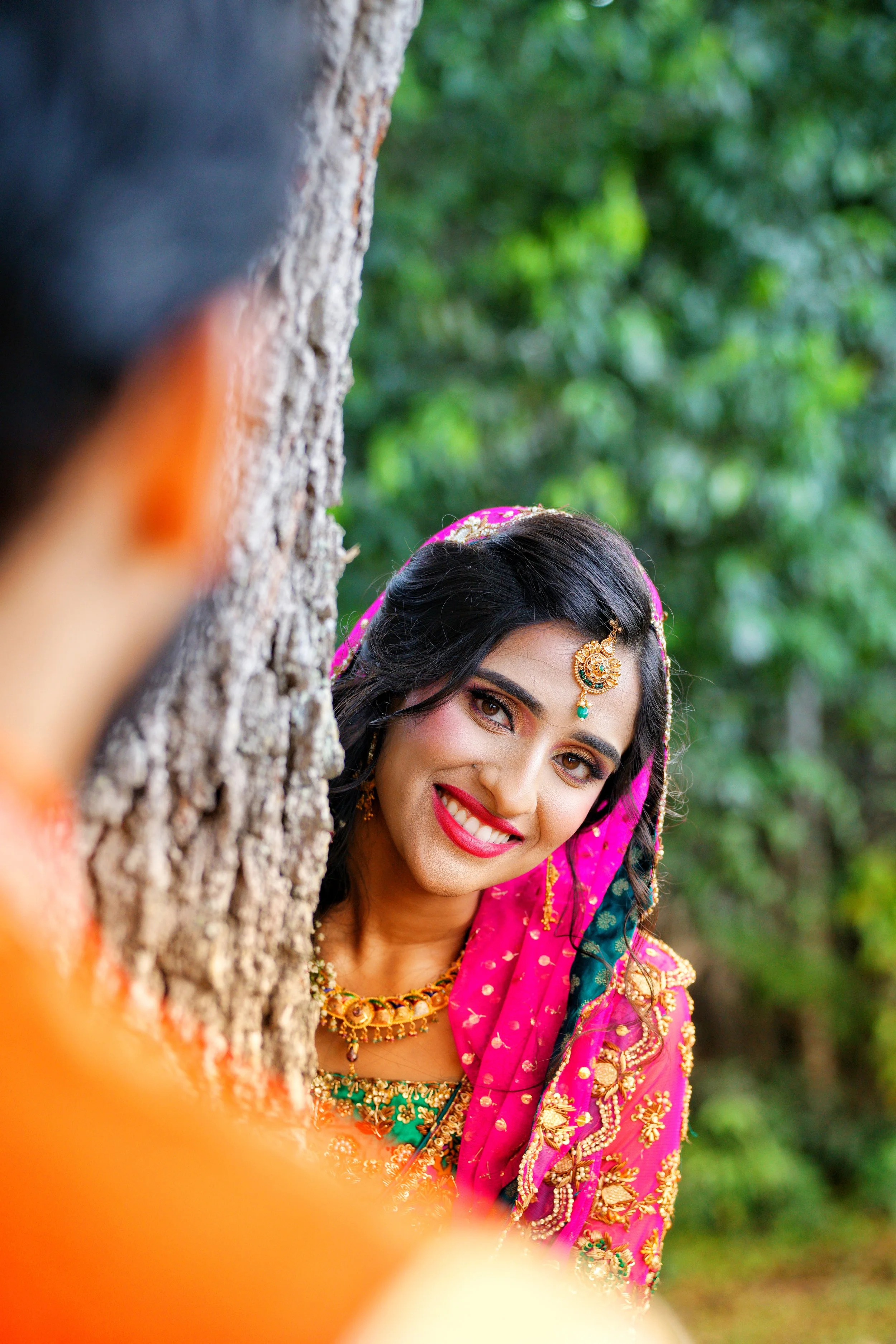 A woman dressed in a traditional pink and green Indian saree with gold embroidery, smiling and looking out from behind a tree in an outdoor setting with green leaves in the background.