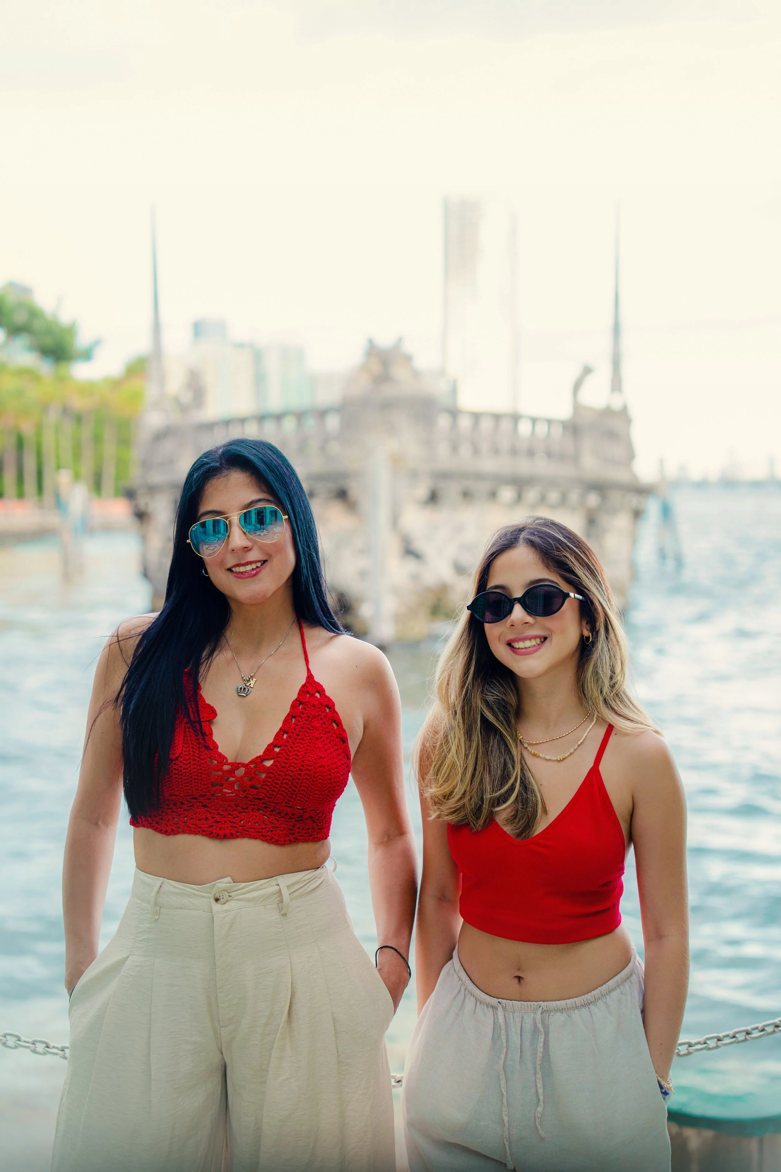Two women smiling at the camera, standing near the water with a boat and cityscape in the background, both wearing sunglasses and summer clothing.