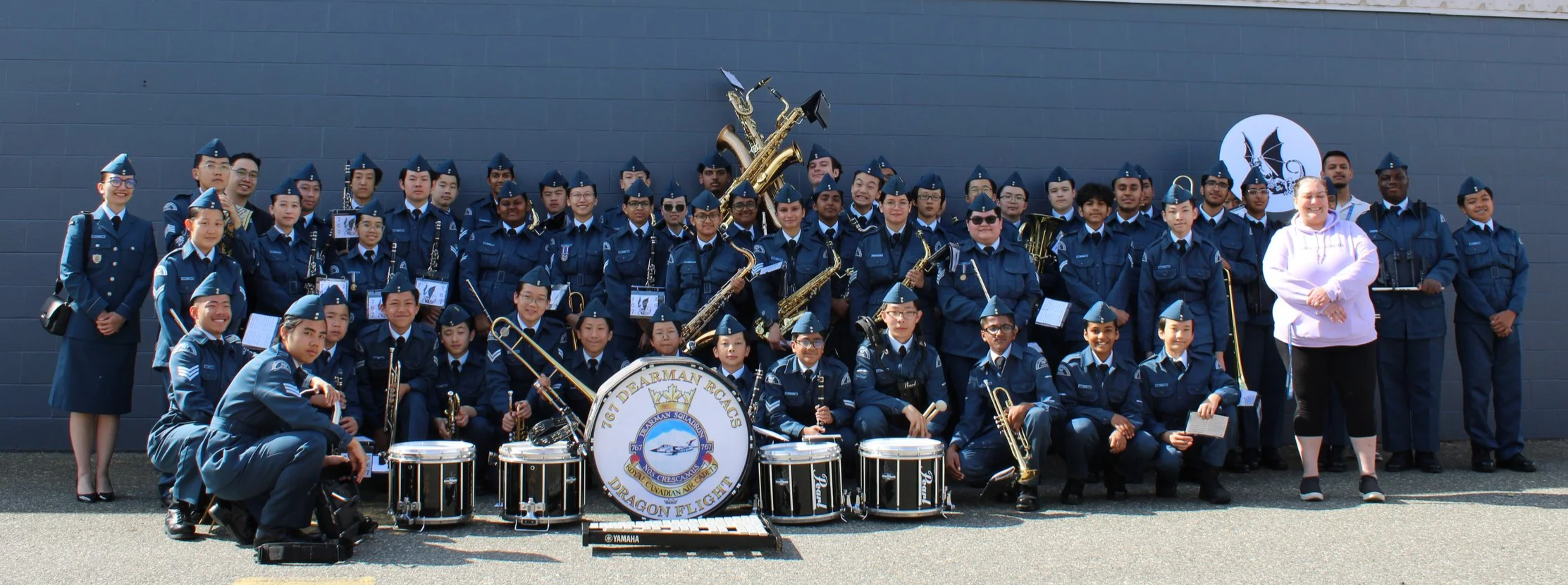 Group of people in uniform posing with musical instruments in front of a dark wall, with a woman in a white hoodie standing to the right.