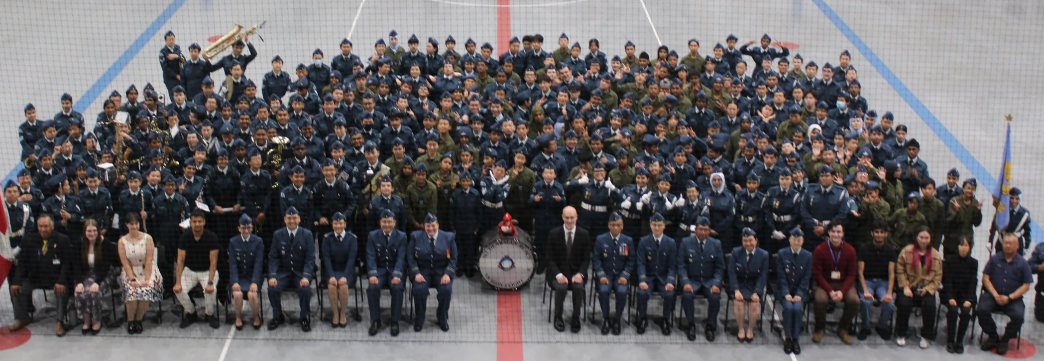 Group photo of people gathered on an indoor ice rink, including individuals in uniforms, casual clothing, and someone with musical instruments, with flags on either side of the group.