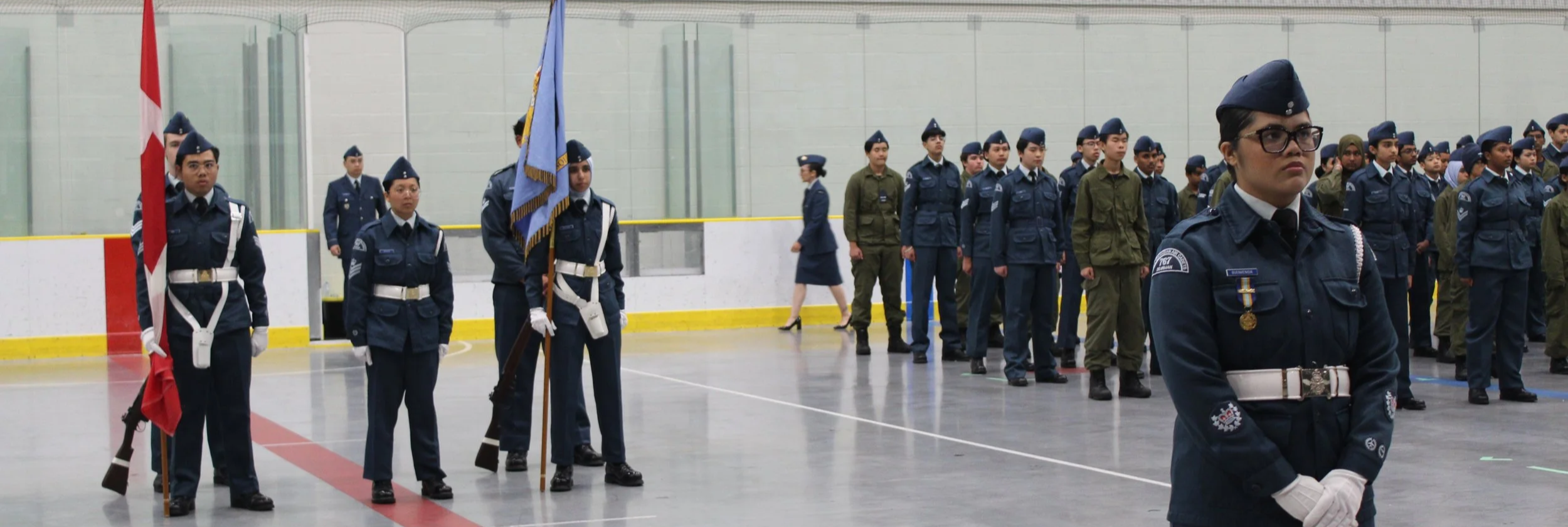 A group of uniformed individuals standing in formation inside an indoor arena, with a person in front holding a Canadian flag and others in military or police uniforms.