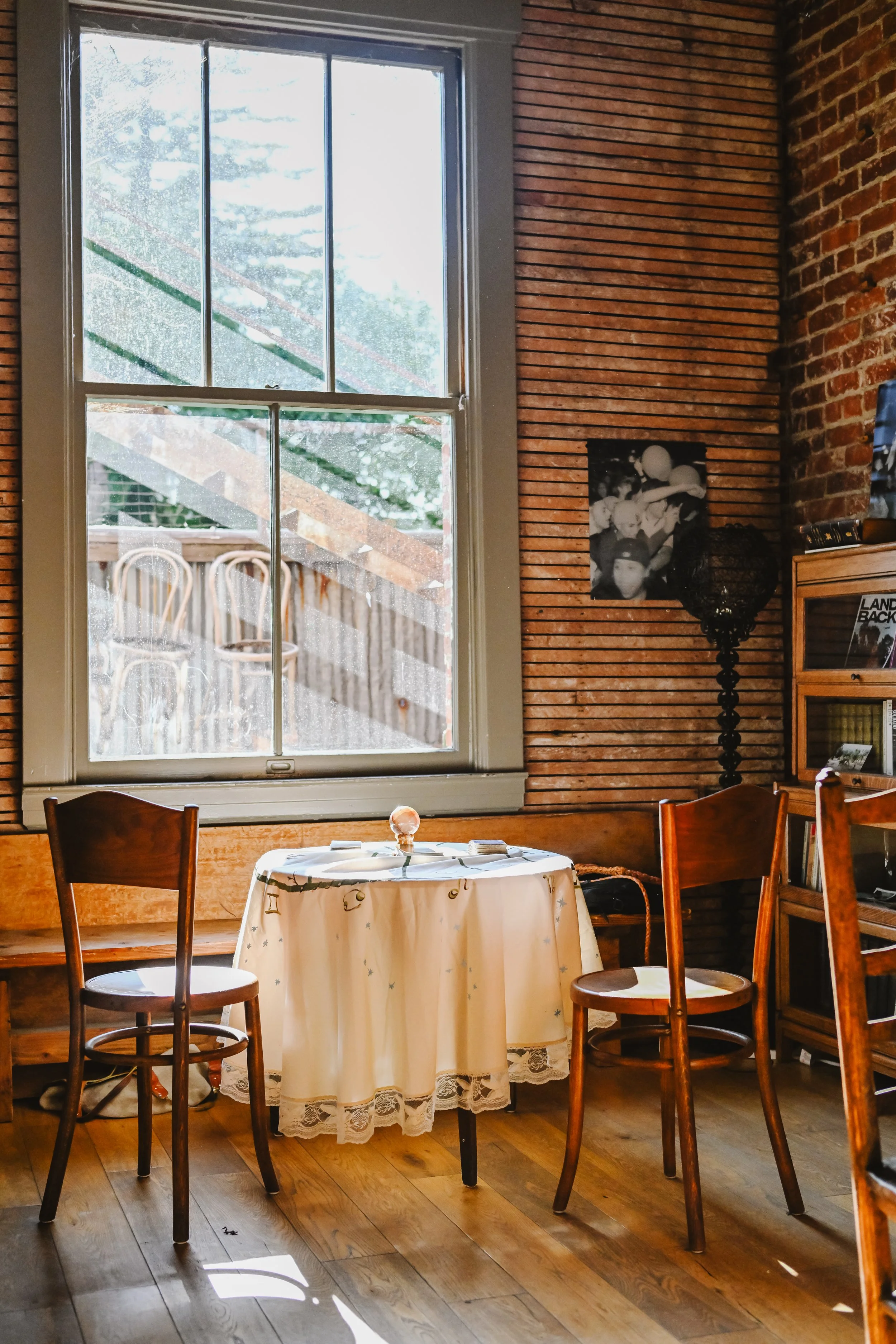 A cozy indoor scene with a round table covered with a white tablecloth decorated with stars and moons, set near a large window with rain droplets. There are wooden chairs around the table and a brick and wooden wall with photo prints and decor behind it.