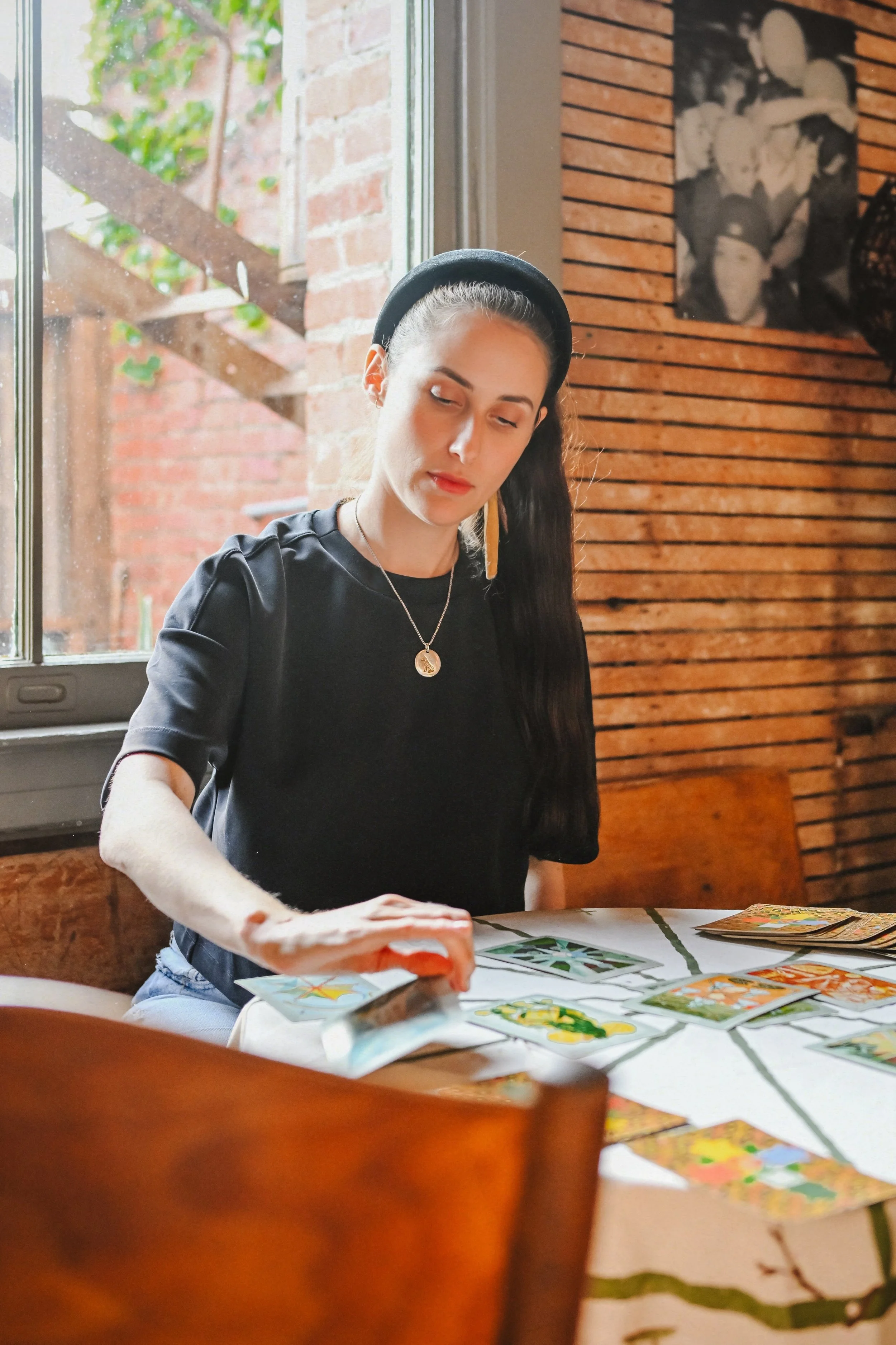 A woman with long dark hair, wearing a black headband, black top, and necklace, sits at a table indoors with tarot cards laid out. She appears to be shuffling or arranging the cards, with a window showing an outside brick wall and greenery behind her.