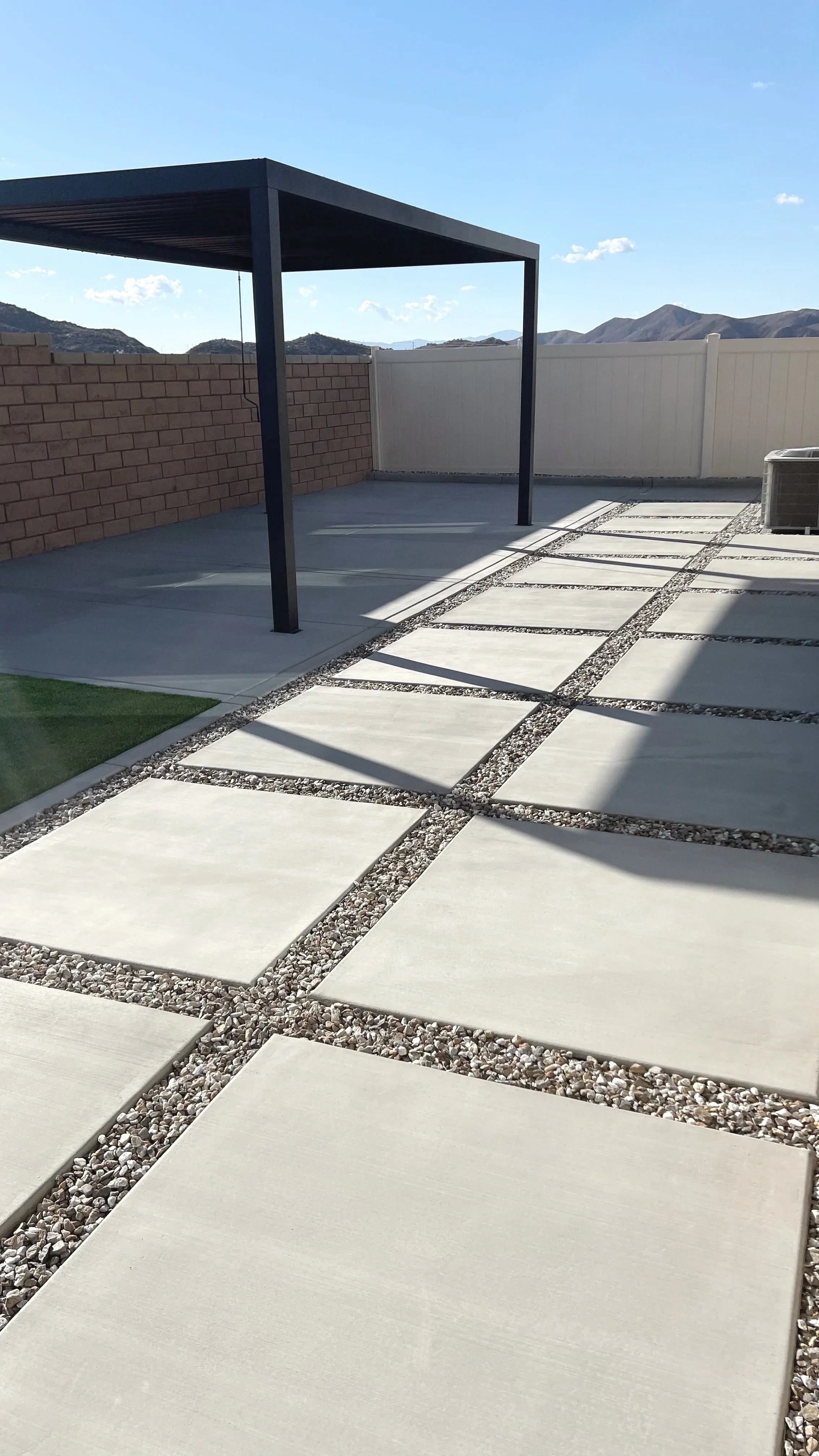 Backyard patio with concrete pavers and gravel in between, a small patch of artificial turf, a shaded structure with black metal posts, a brick wall, a white fence, and mountains in the background.