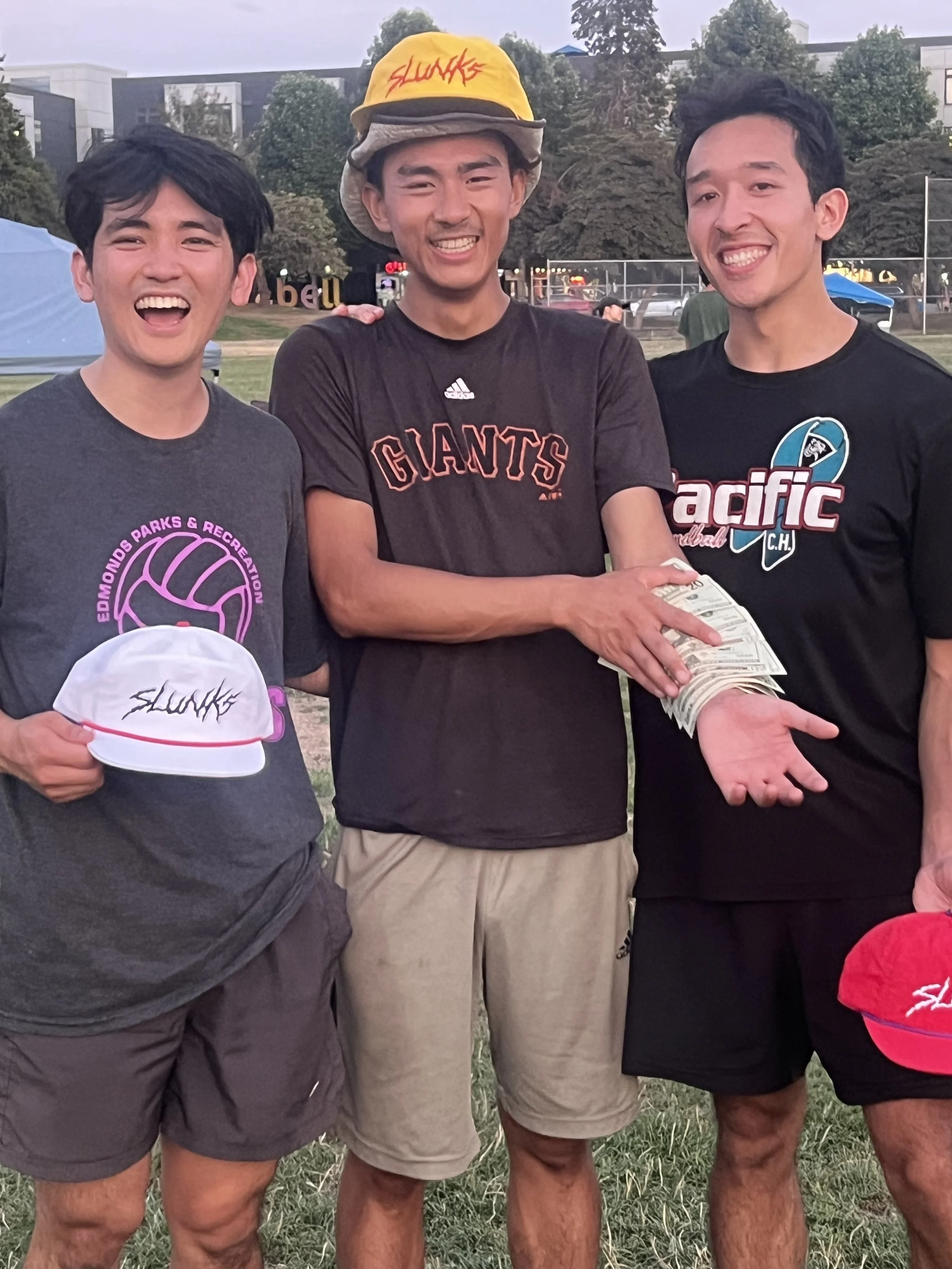 Three young men standing outdoors at a park, smiling and holding baseball caps and cash. One man in the middle wears a yellow bucket hat and a black shirt with 'GIANTS' written on it. The man on the left holds a white cap with 'Slurve' written on it and wears a gray shirt with a pink and purple volleyball design. The man on the right wears a black shirt with 'Pacific' written on it.