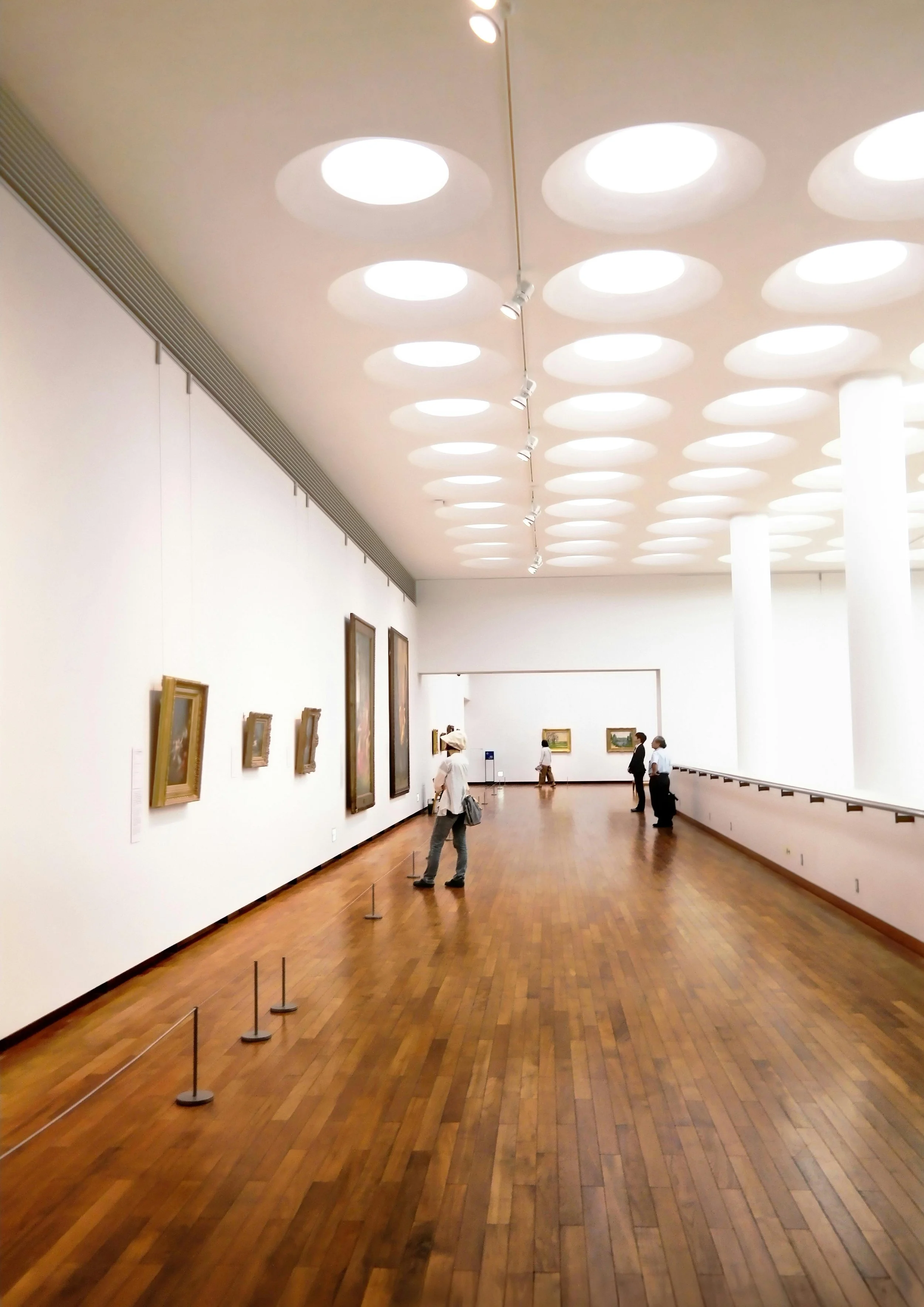 Interior of an art gallery with white walls, wooden floor, several paintings on display, and a few visitors looking at the artwork.