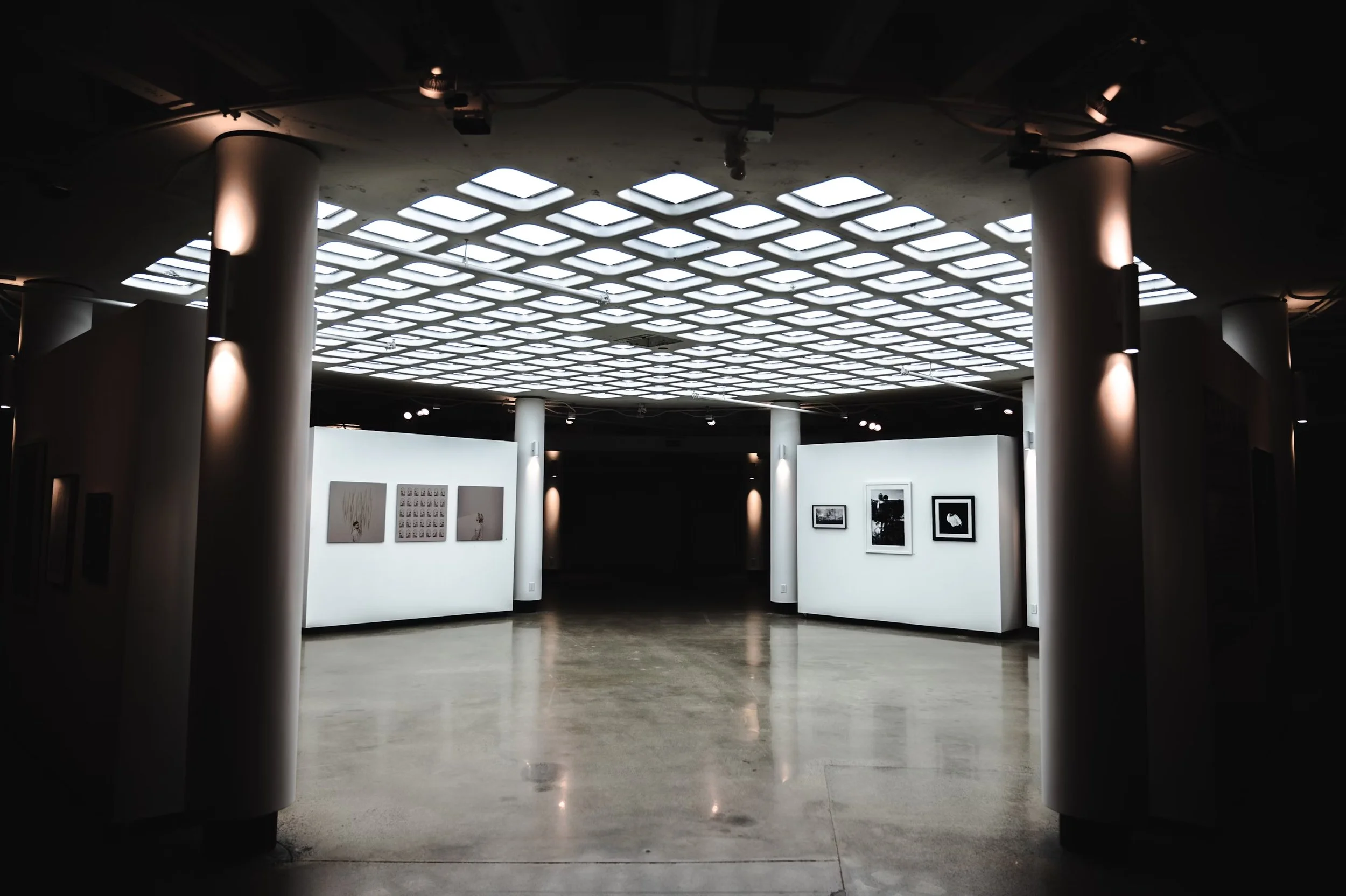 Interior of an art gallery with white walls displaying framed artwork and a decorative ceiling with grid-patterned lighting.