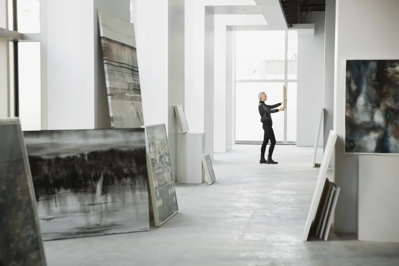 Person viewing artwork in a modern art gallery with paintings on display and large windows letting in natural light.