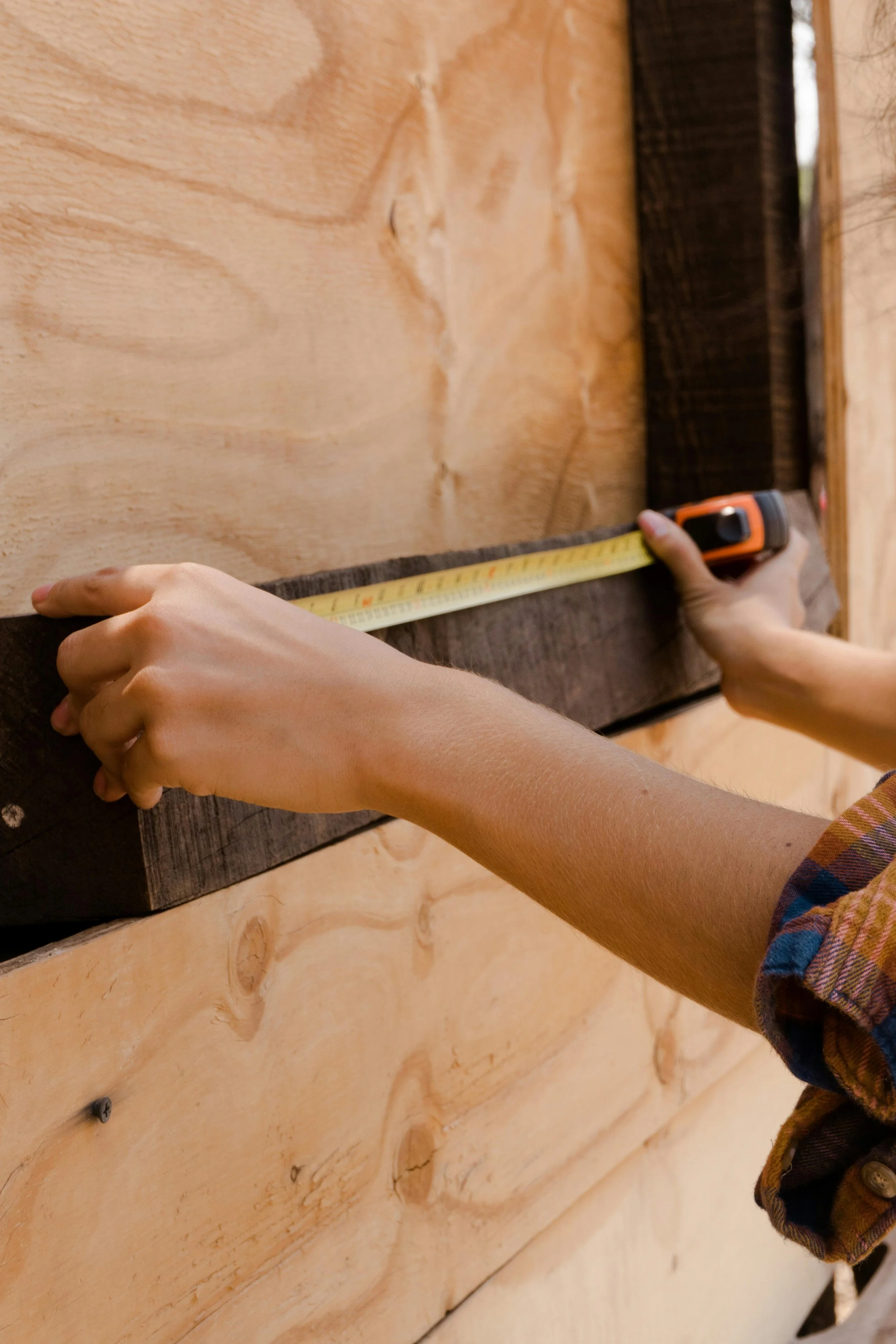 An art expert is measuring a piece of dark wood with a tape measure against a light wood wall, for art packaging and crating.