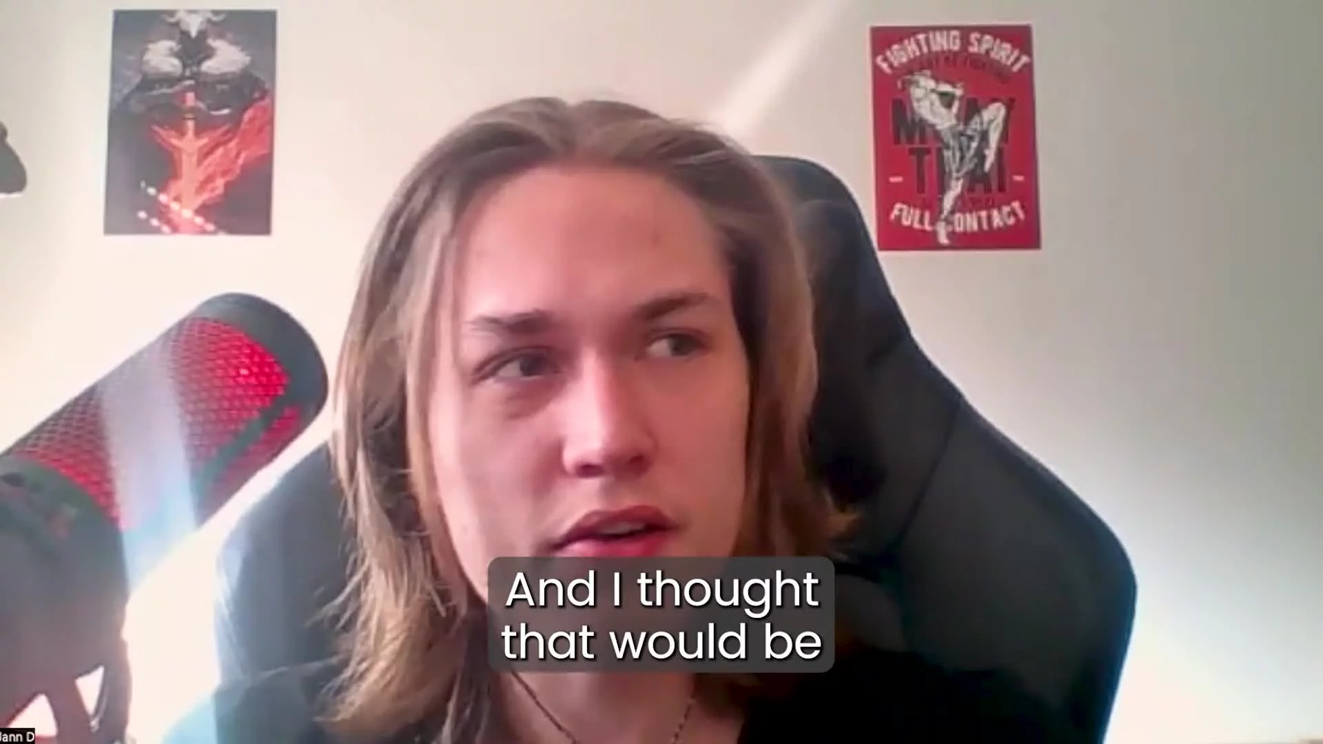 A young person with long hair sitting in front of a microphone, with posters on the wall behind them, one showing a red fire and black figure, and another with martial arts imagery and text about fighting spirit.
