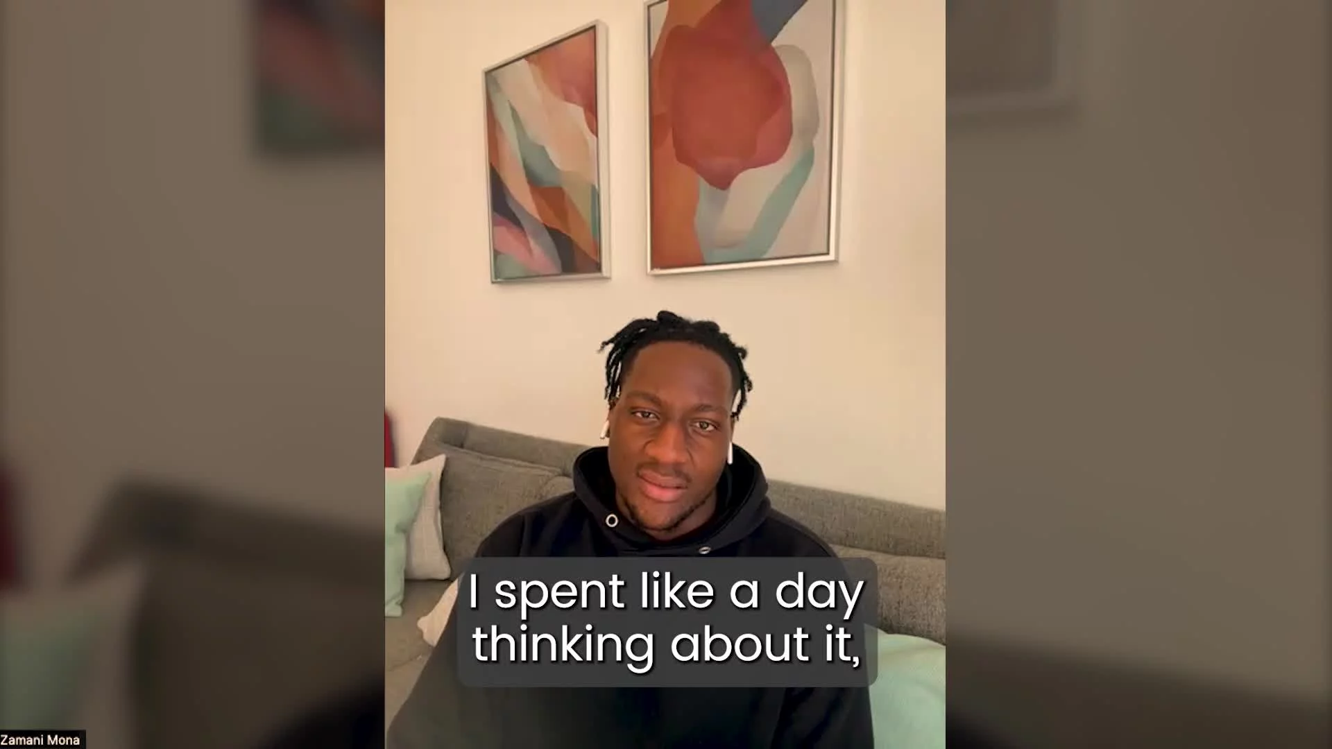A man with dreadlocks wearing a hoodie and wireless earbuds, sitting on a sofa in a living room, with abstract art on the wall behind him. Subtitles read: 'I spent like a day thinking about it,'.
