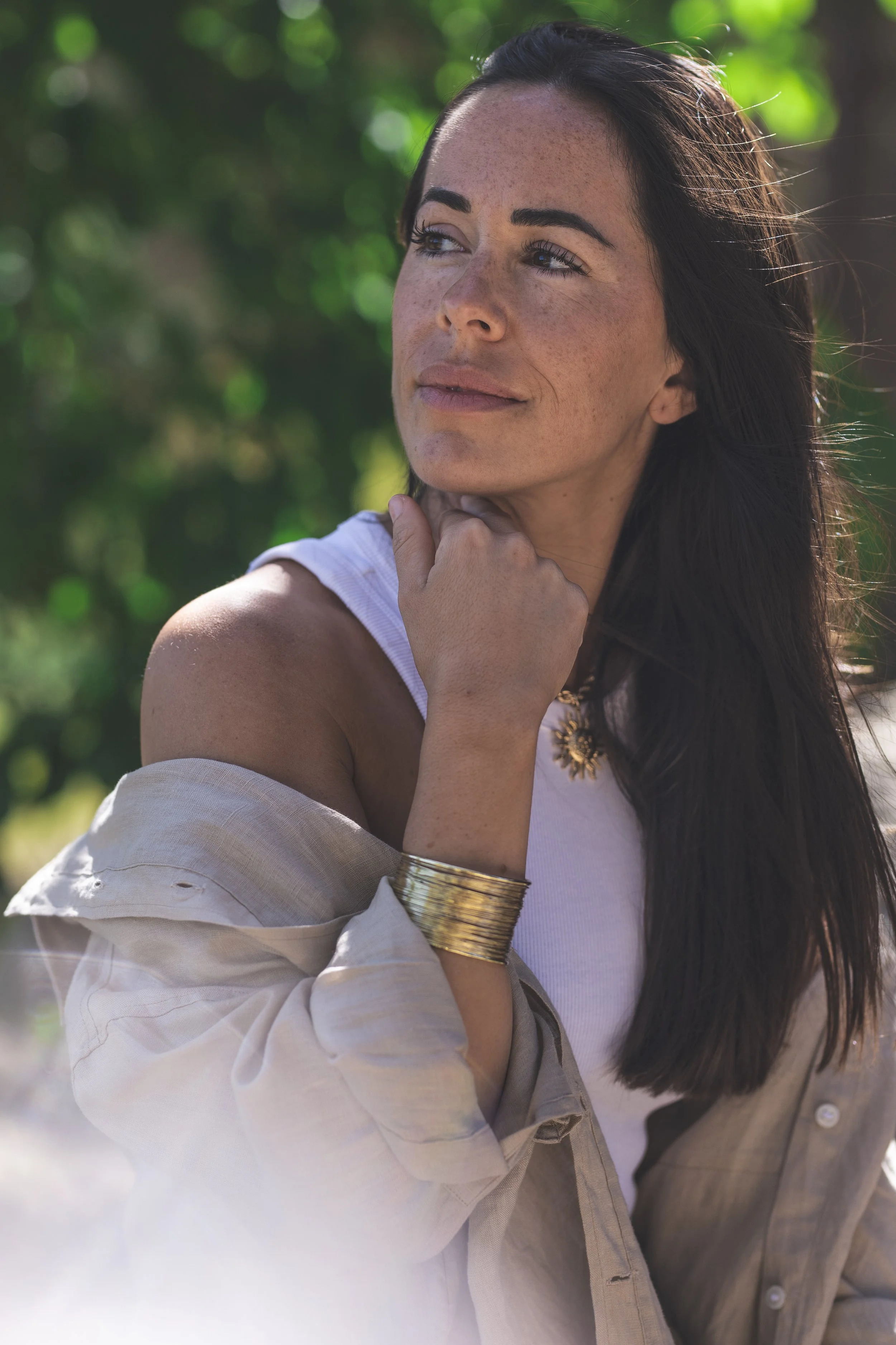Joven mujer de cabello largo, oscuro y con manchas de pecas, mirando hacia arriba y sonriendo, en un entorno al aire libre con fondo de árboles y naturaleza.