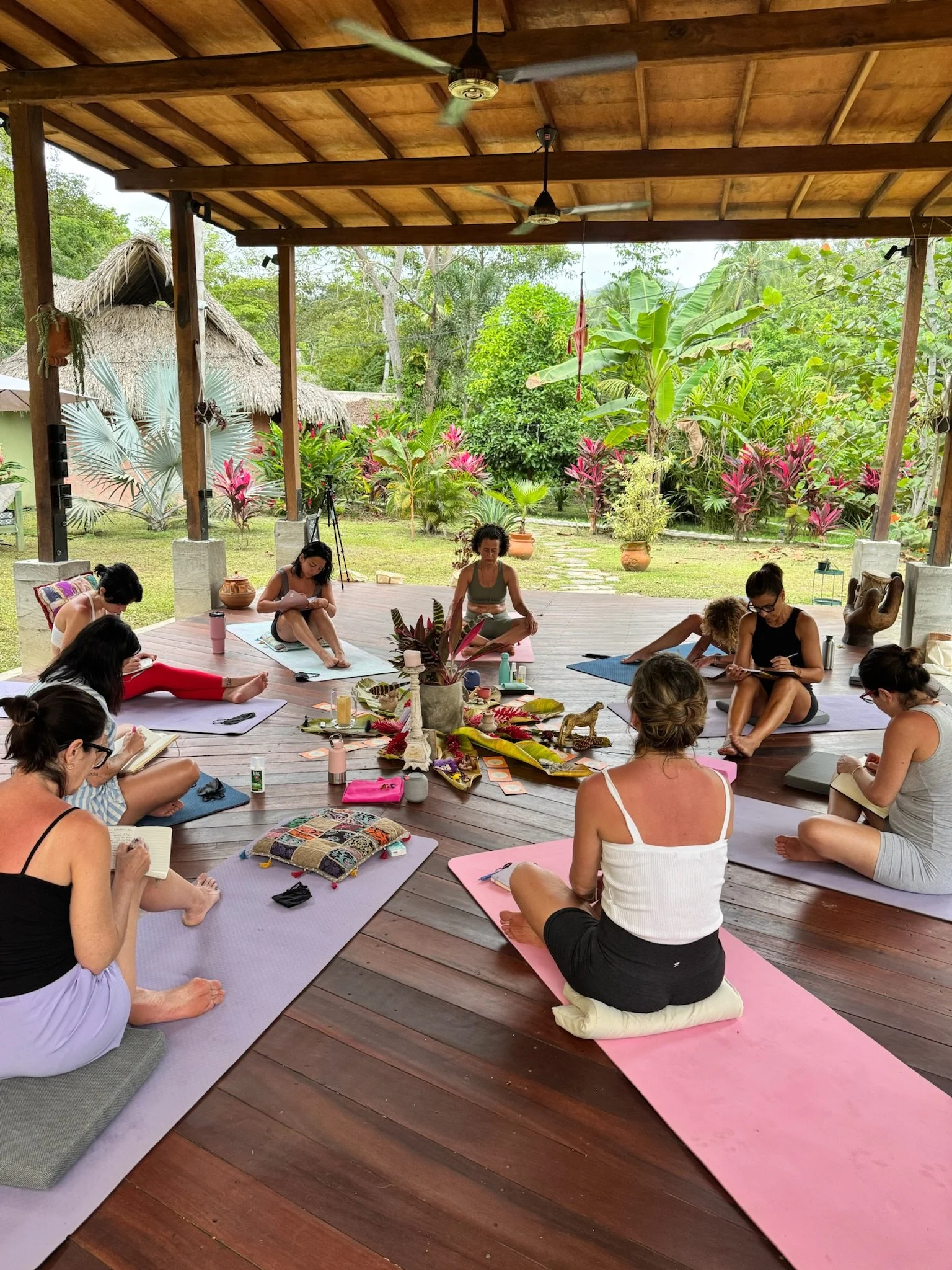 Un grupo de personas participando en una clase de yoga al aire libre bajo un pórtico de madera, rodeadas de vegetación tropical y plantas.