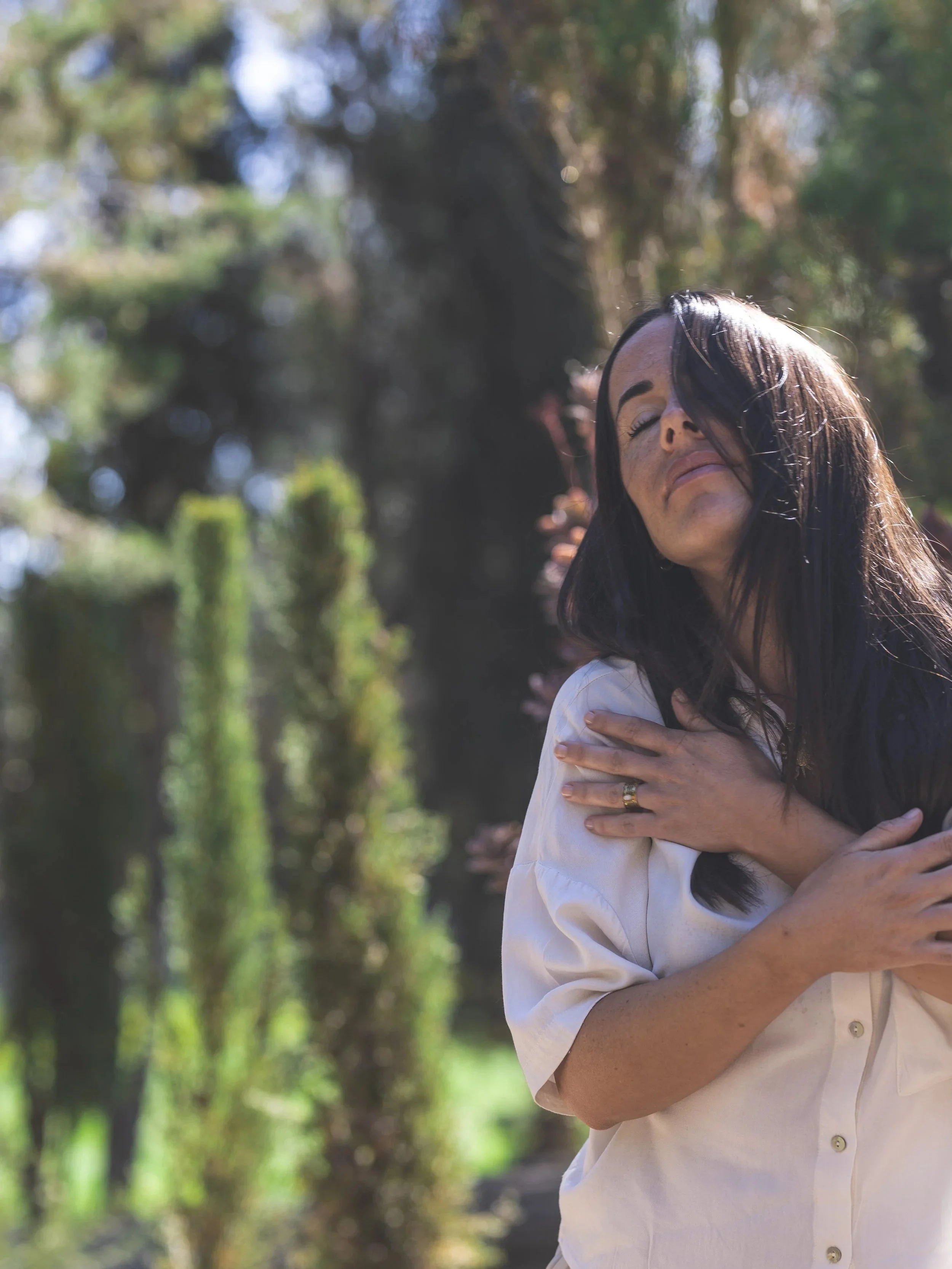 Mujer con cabello negro y piel clara, con expresión de emotividad, abrazándose a sí misma en un ambiente natural con árboles y luz solar difusa.