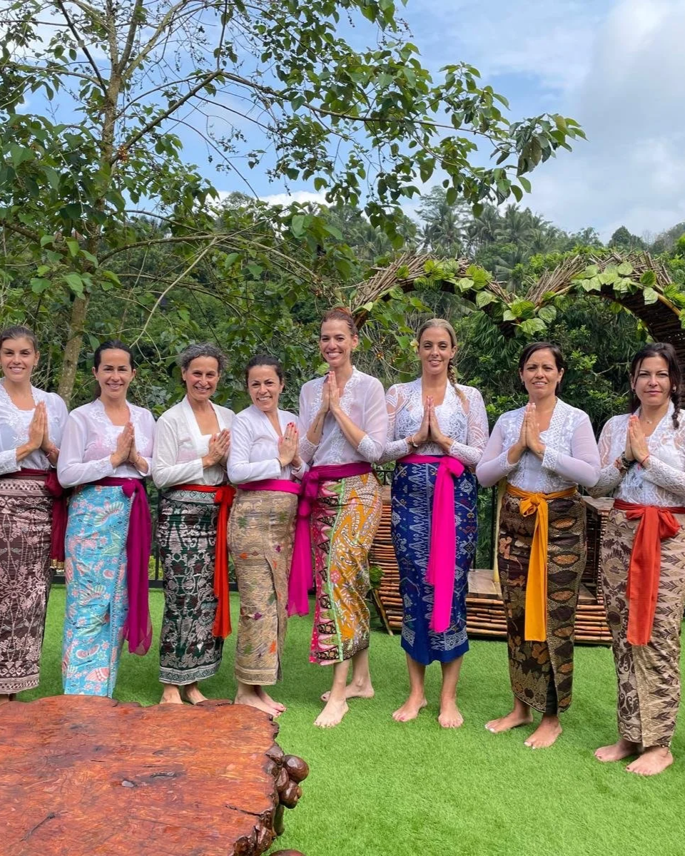 Nueve mujeres vestidas con ropa tradicional, con sus manos juntadas en señal de saludo o oración, posando en un jardín con vegetación y un arco decorativo en el fondo.