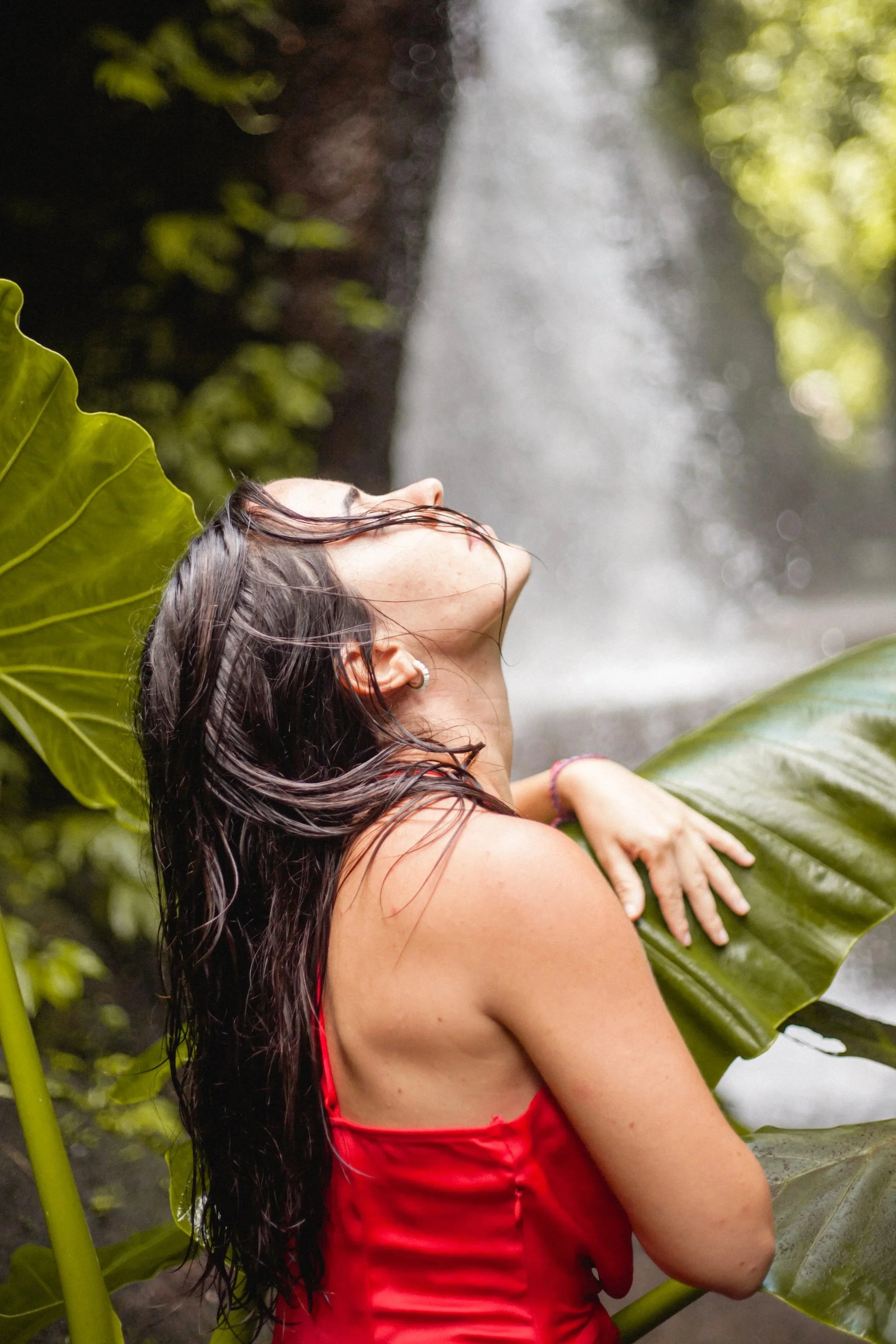 Mujer con cabello largo y oscuro, de piel clara, con ojos cerrados, viste un vestido rojo, rodeada de grandes hojas verdes y en un fondo de una cascada en un entorno natural selvático.