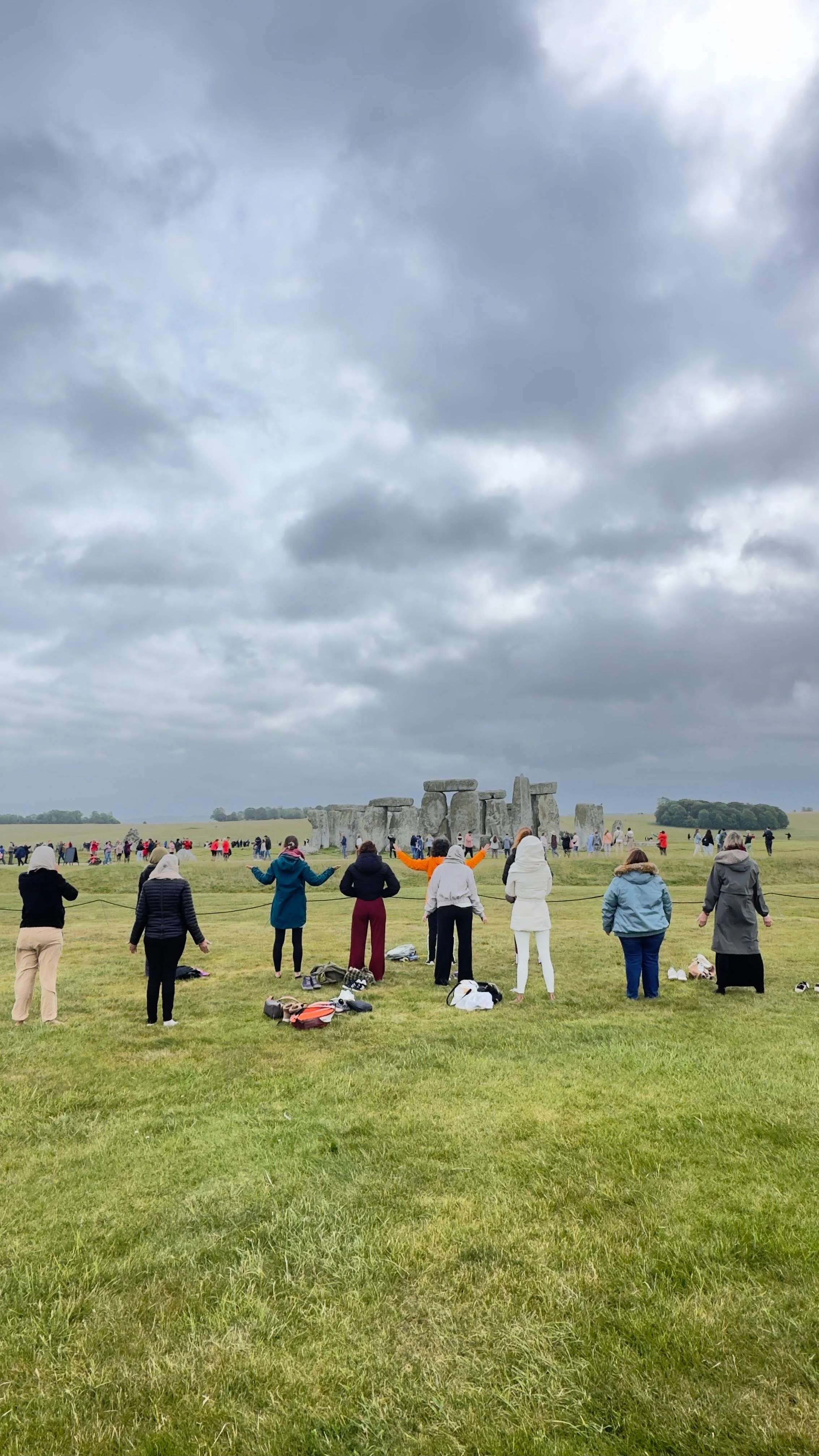 Personas observando el monumento de Stonehenge en un día nublado, en un campo abierto con cierta hierba, muchas mochilas y ropa de abrigo.