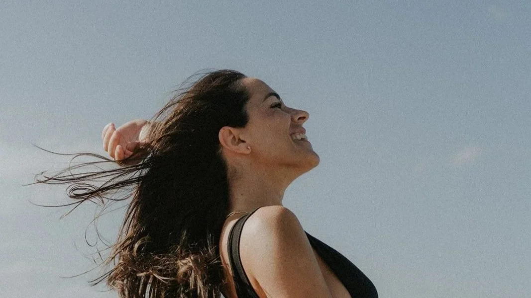 Mujer sonriendo con cabello al viento en un día soleado y cielo despejado.