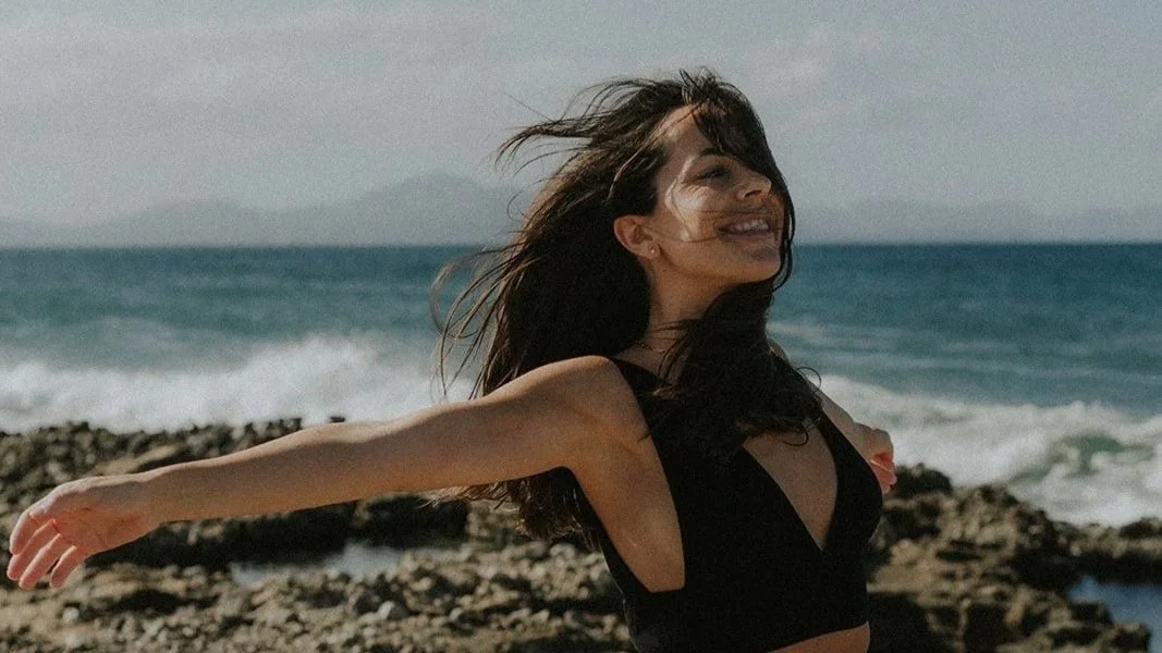 Mujer sonriendo en la playa con cabello al viento, fondo mar y montañas.