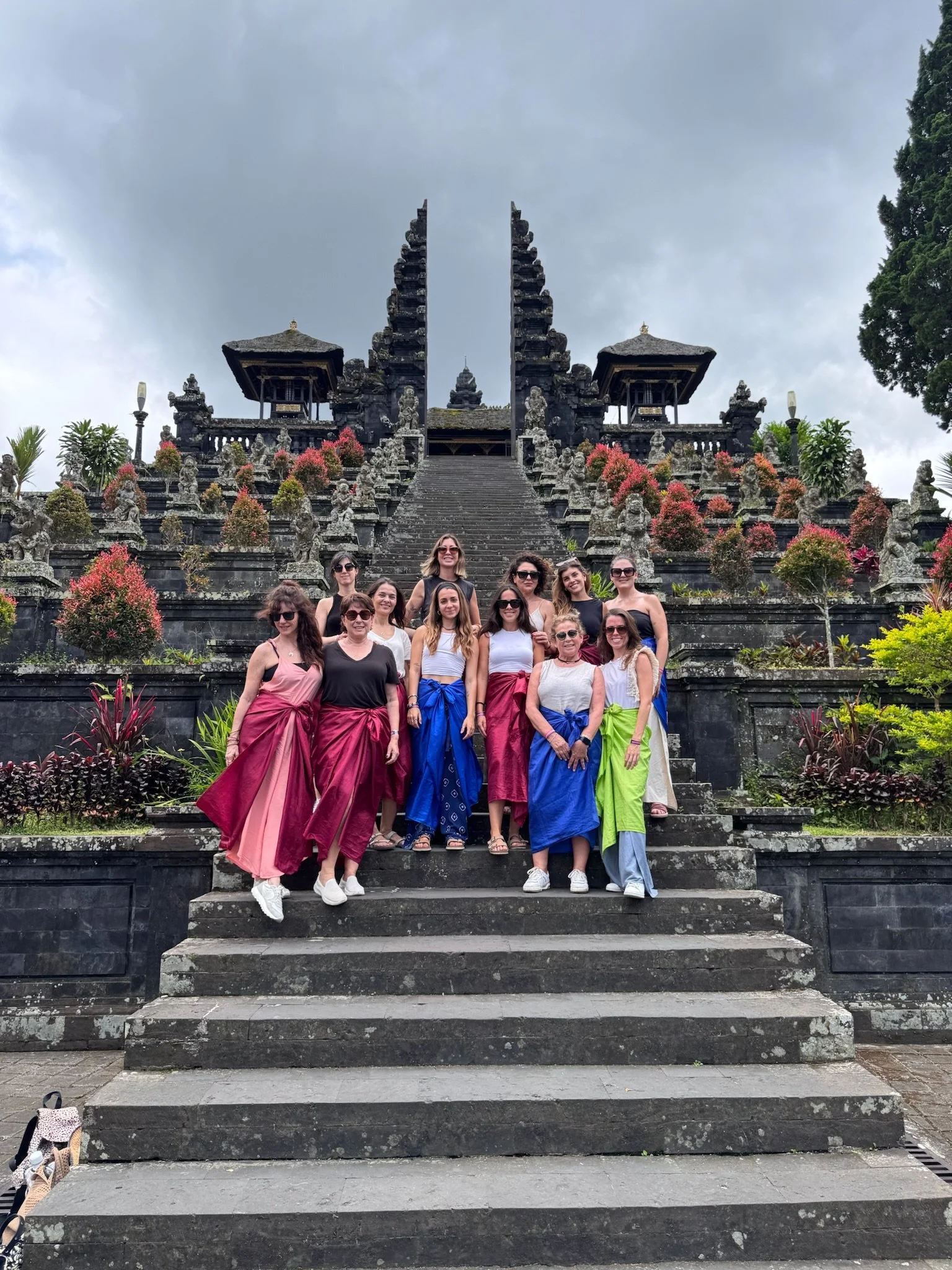 Un grupo de personas posando en la entrada de un templo o monumento en Bali, con escaleras y estatuas, rodeado de plantas y árboles, en un día nublado.