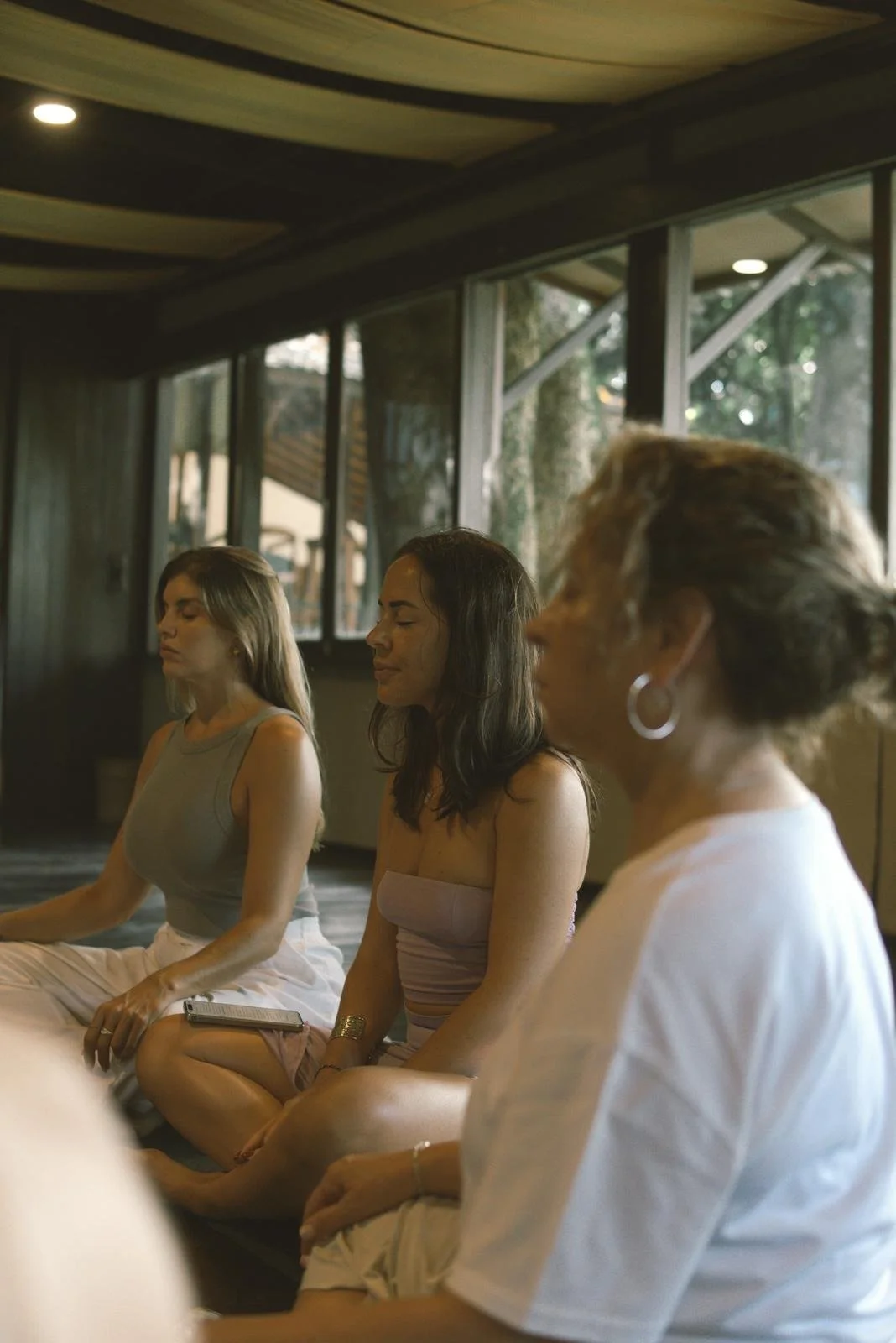 Tres mujeres en posición de meditación, sentadas con las piernas cruzadas, con los ojos cerrados en una habitación con grandes ventanas y luz natural.
