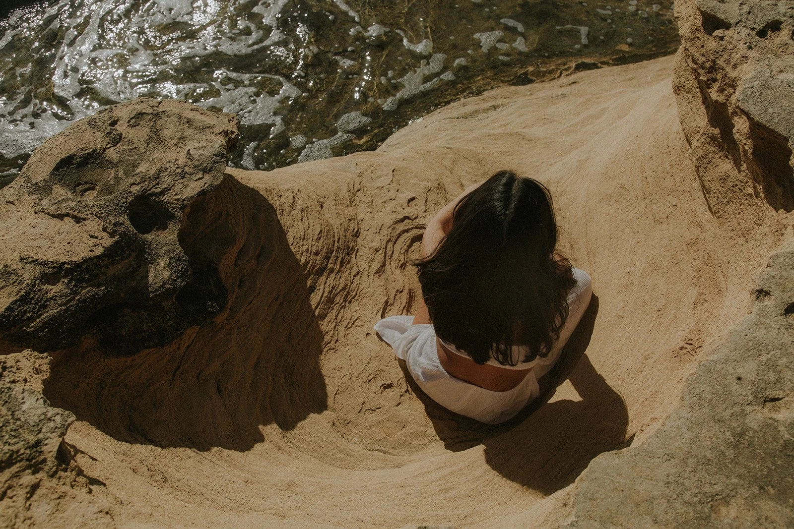 Una mujer con cabello oscuro y vestimenta blanca sentada en una roca en una zona de playa, observando hacia abajo, rodeada de formaciones rocosas y agua en el fondo.