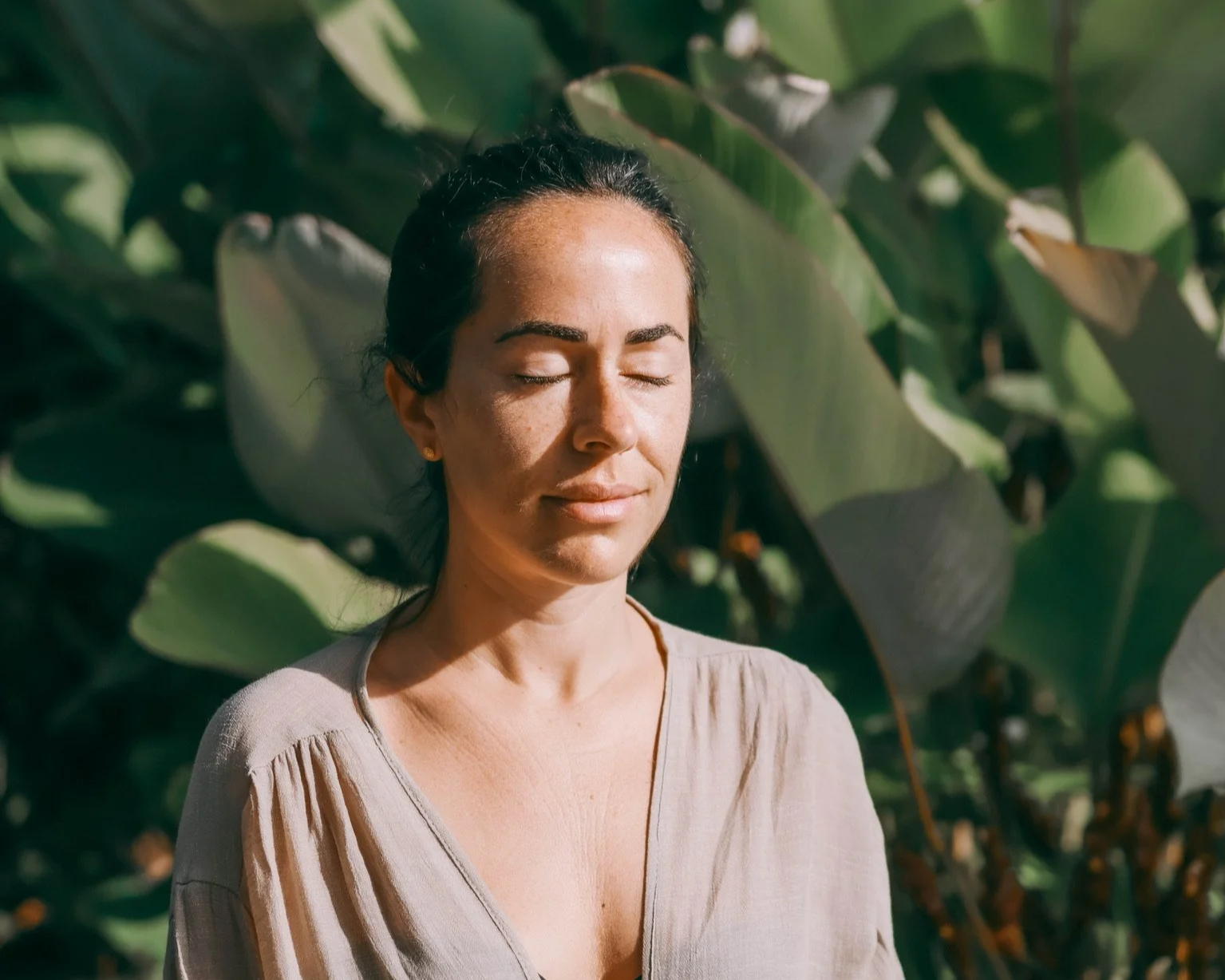 Mujer con cabello oscuro y piel clara, con ojos cerrados, en un entorno con plantas verdes.