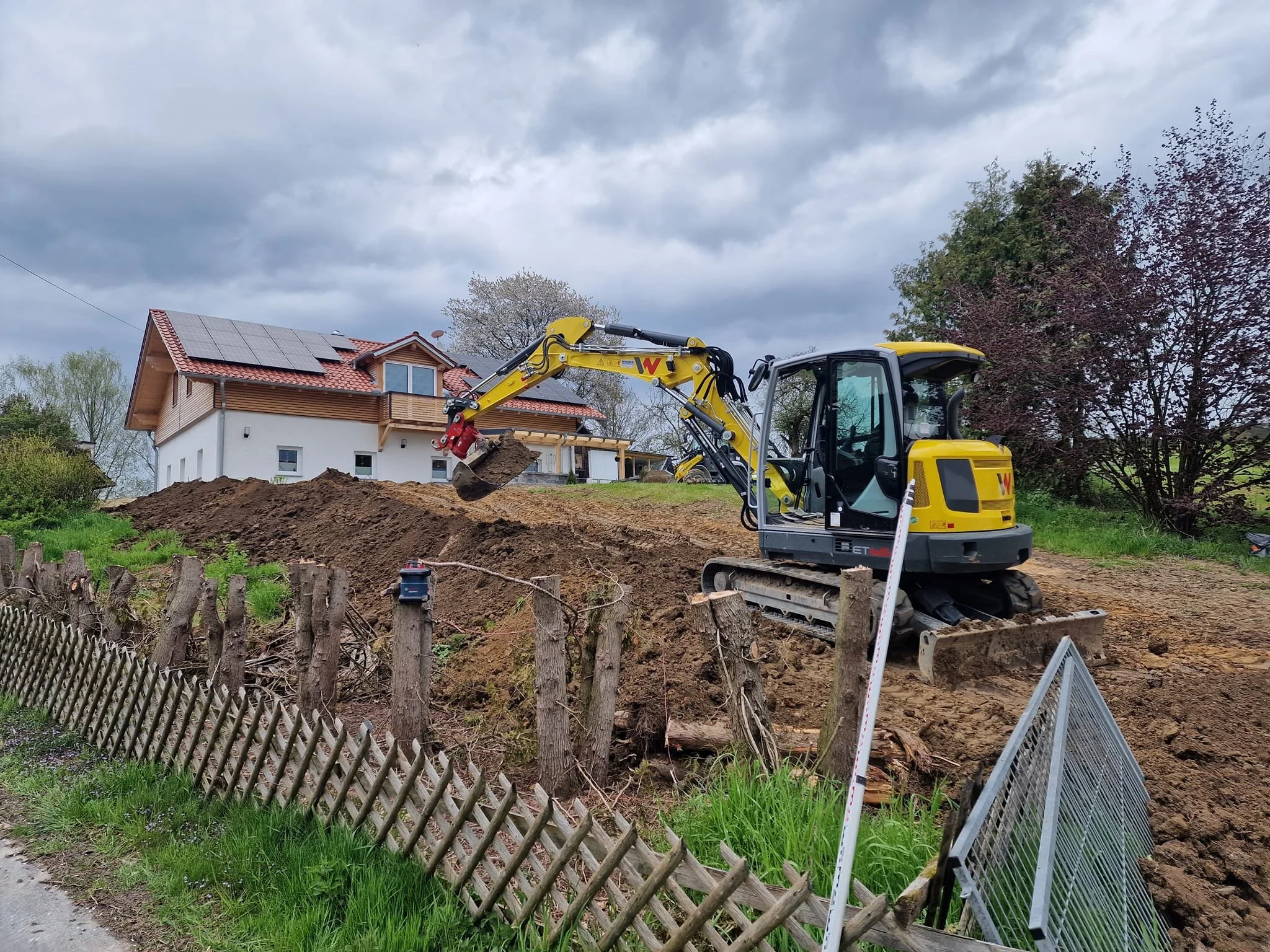 Bagger arbeitet auf einer Baustelle vor einem Haus mit Solardach, umgeben von Bäumen und einem Zaun.