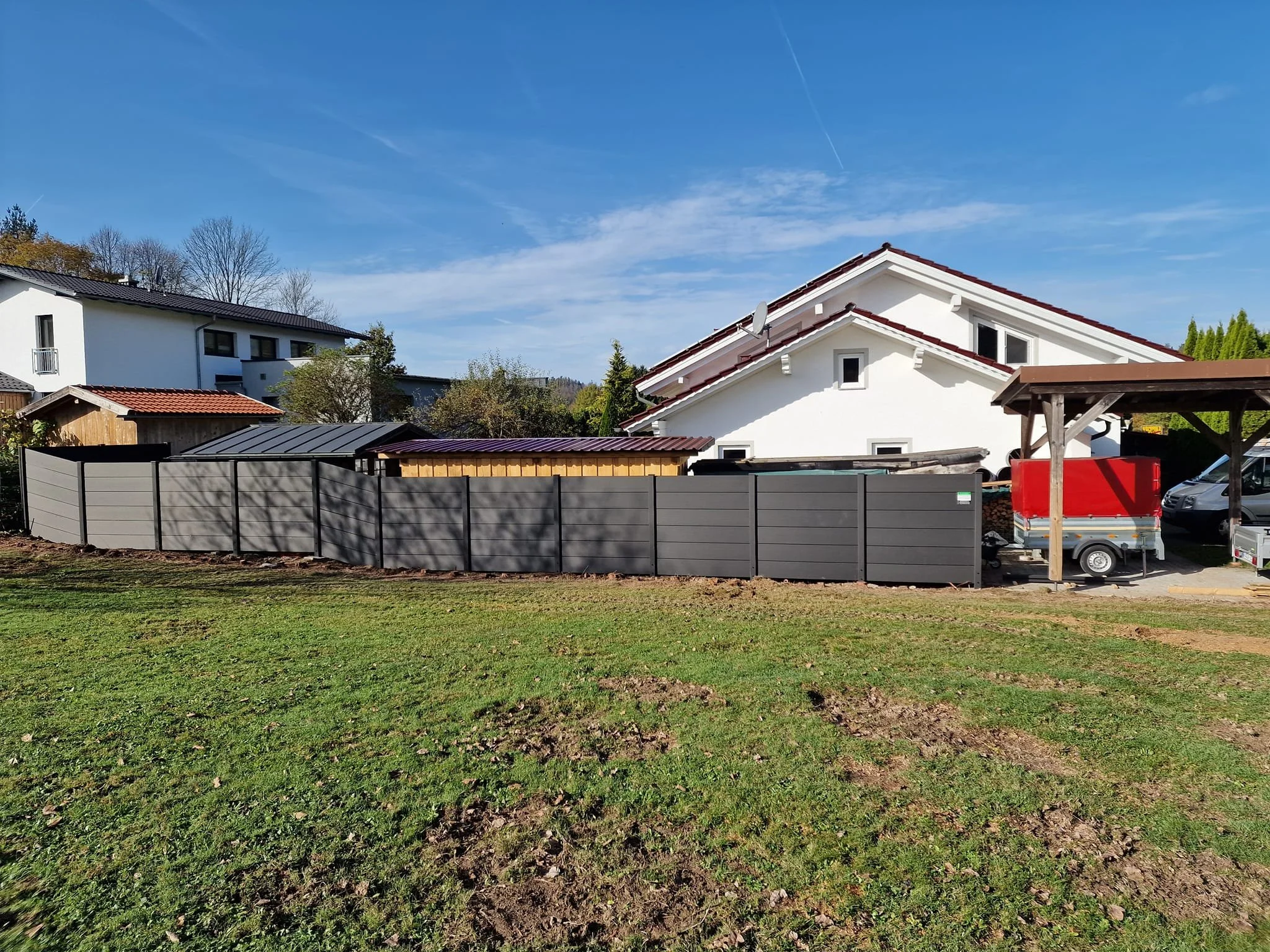 Garten mit begrüntem Rasen, metallischer Sichtschutzzaun, weißes Haus mit Dachgauben und Satellitenschüssel, Carport mit LKW und Autos, blauer Himmel mit Wolken.