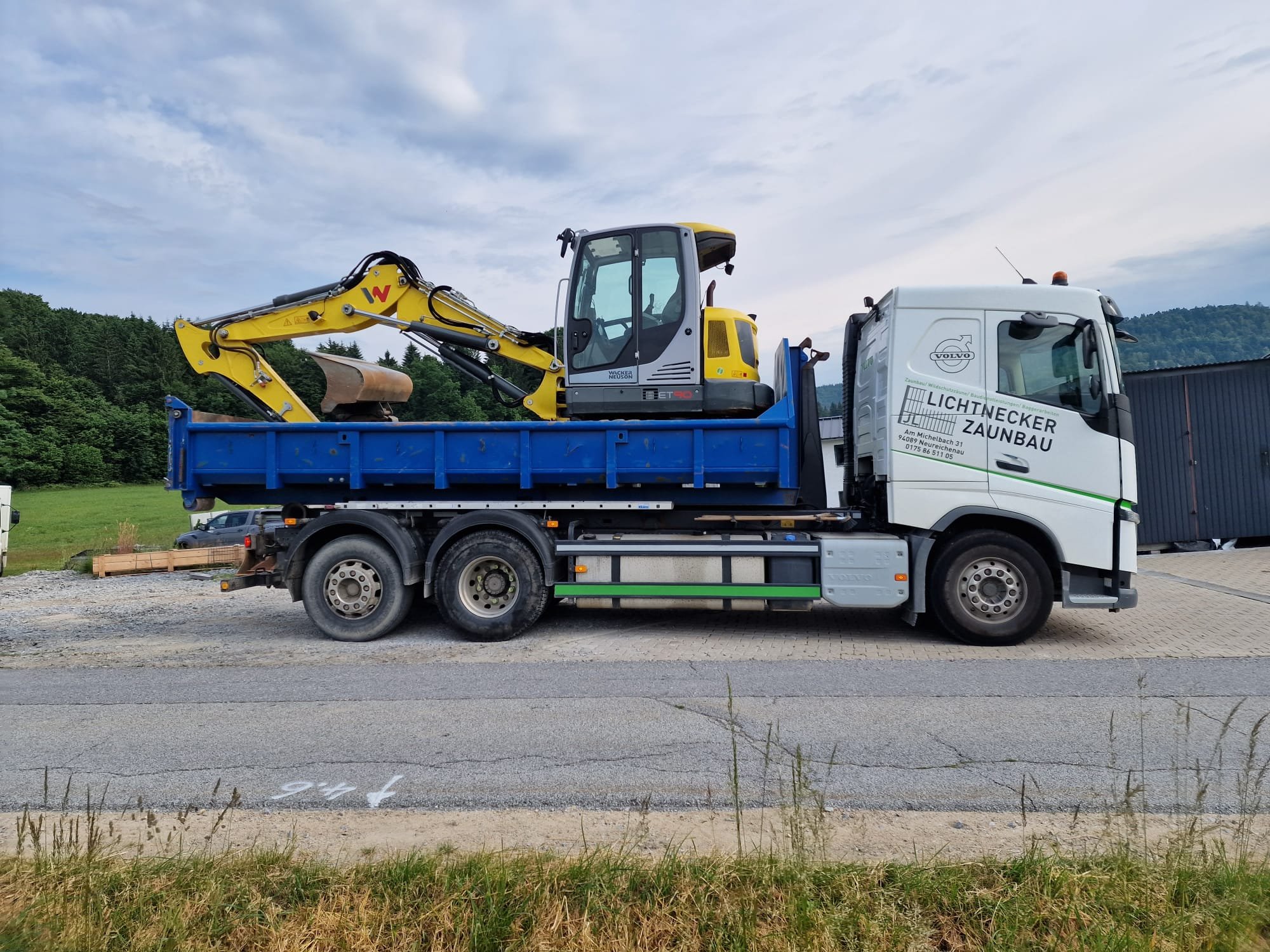 LKW mit Bagger auf einer Baustelle, grüner Hügel im Hintergrund.