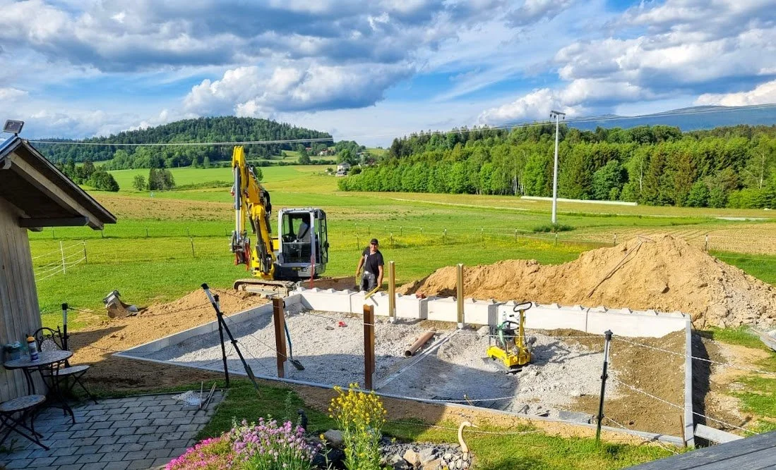 Baustelle im ländlichen Gebiet mit Bagger, Bauarbeiter, Fundamentarbeiten und angrenzende grüne Felder und Wälder unter bewölktem Himmel.