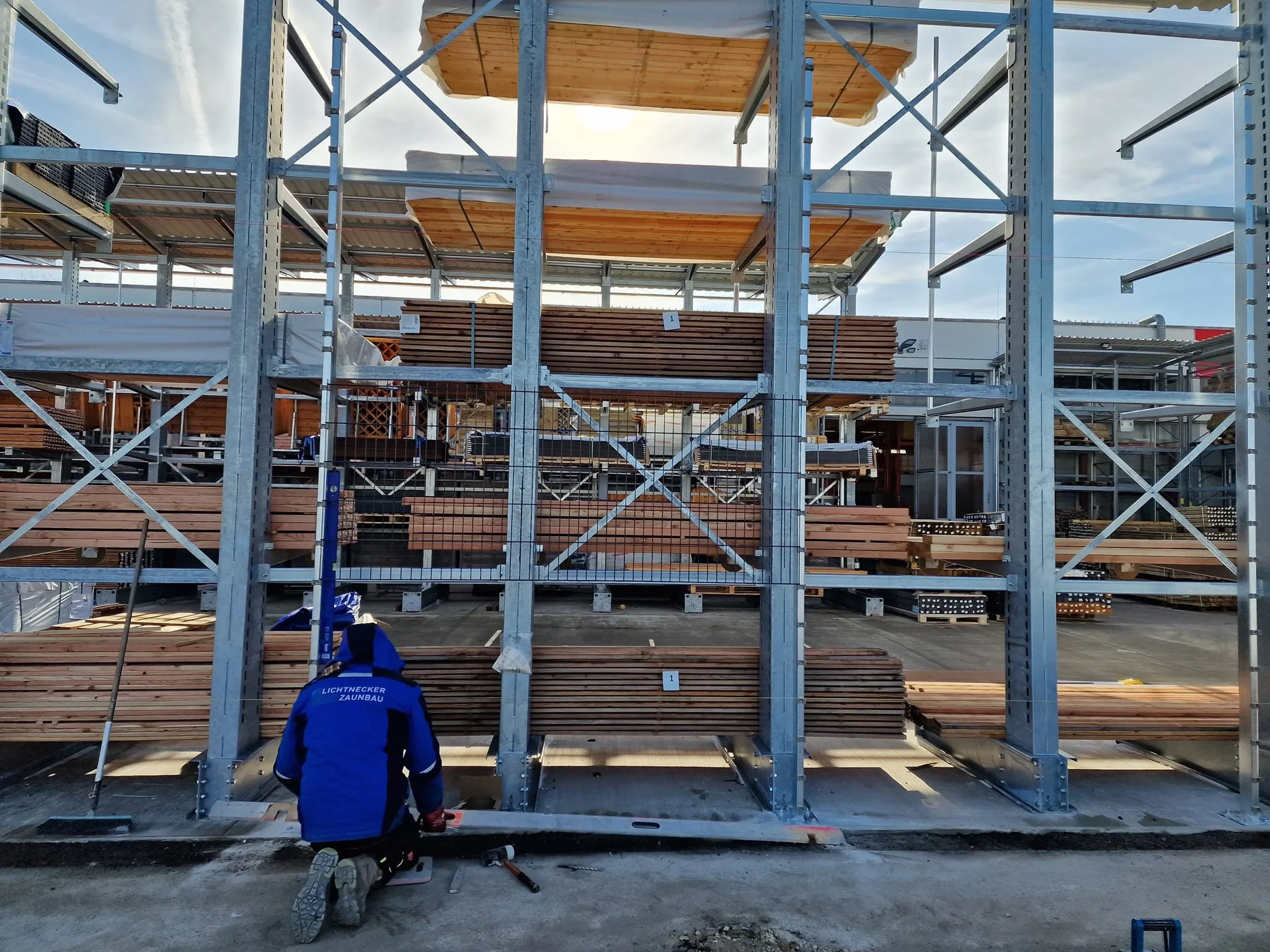 Ein Bauarbeiter in blauer Jacke mit der Aufschrift 'Lichtnecker Zaunbau' arbeitet an einem Gerüst aus Metall und Holz auf einer Baustelle, um ein Gebäude zu errichten.