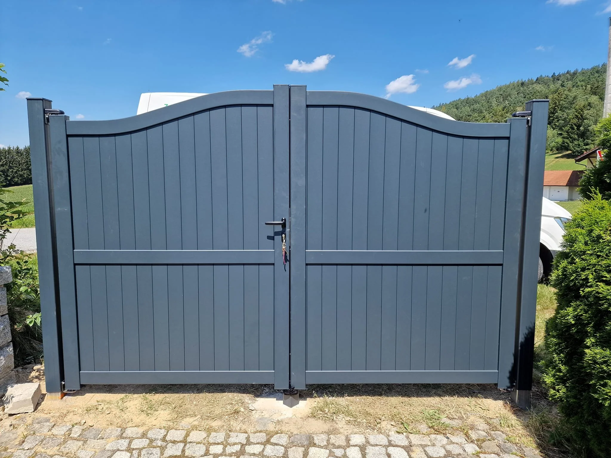 Ein grauer Hoftor-Tor mit ländlicher Umgebung und Garten, blauer Himmel, Wolken, Berge im Hintergrund.