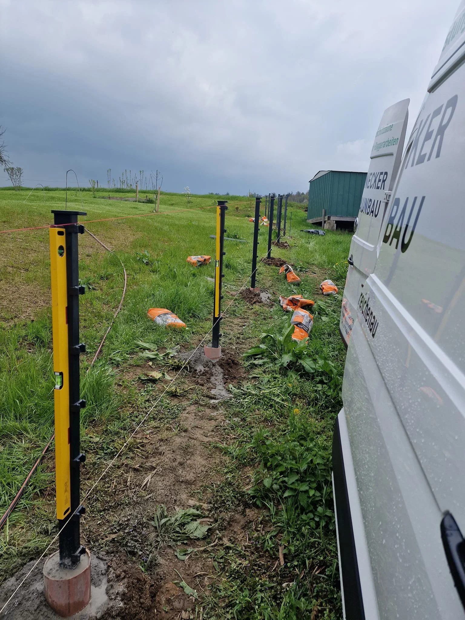 Bodenmarkierung für einen Zaunbau auf einer Wiesenfläche, mit Messwerkzeugen und Absperrungen, bei schlechtem Wetter.