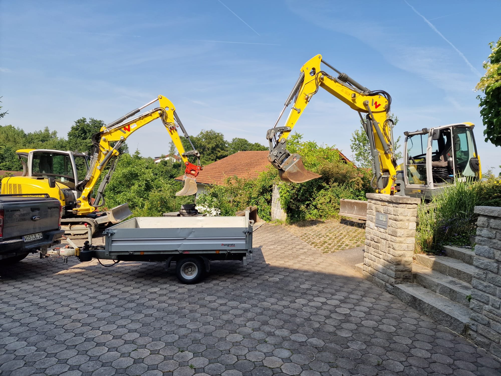 Zwei kleine Baumaschinen mit gelben Armen heben jeweils eine Schaufel aus einer Transportkarre. Sie stehen auf einer gepflasterten Fläche vor einem Haus mit rotem Dach und grünen Bäumen im Hintergrund.