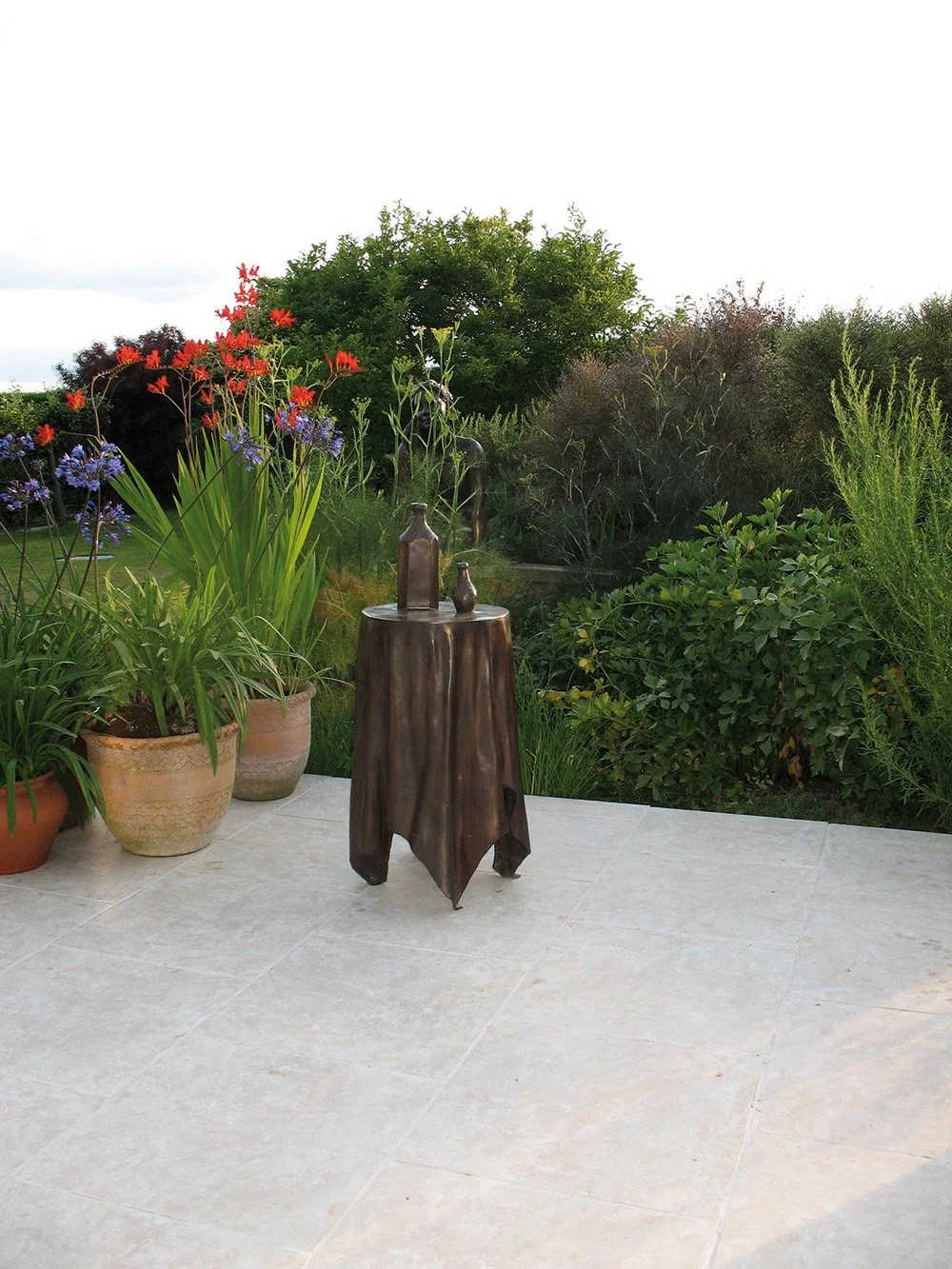 Still Life VI life size bronze sculpture of  circular table tablecloth and bottles in garden setting