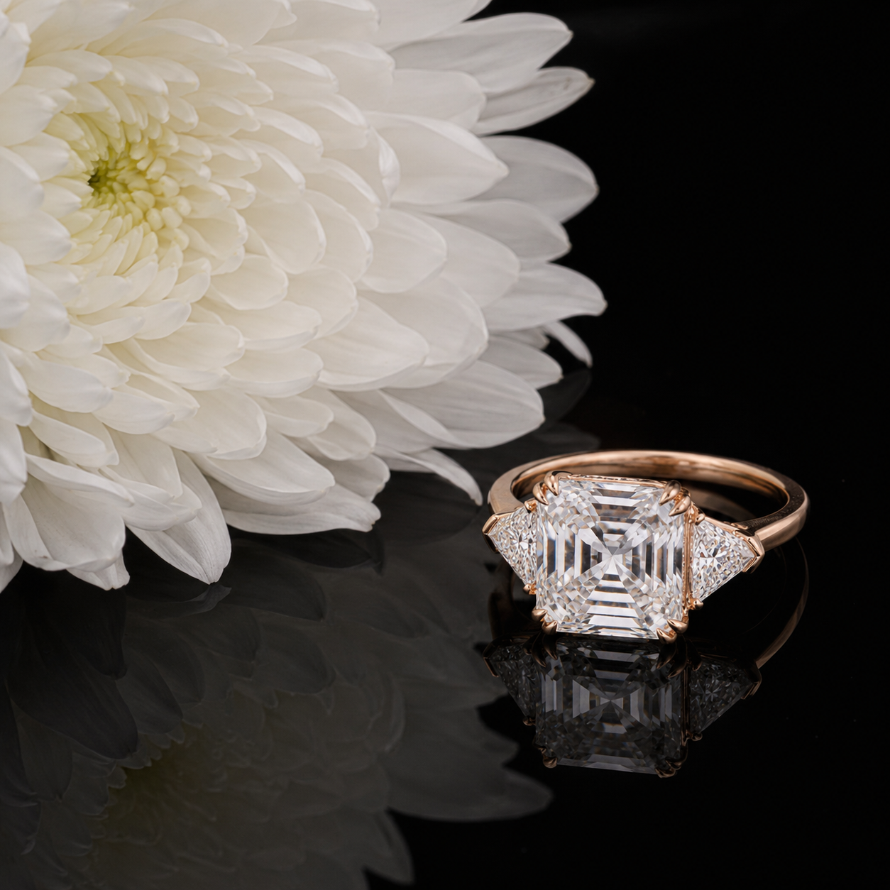 Ein großer, quadratischer Diamant auf einem goldenen Ring neben weißen Blüten auf schwarzem Hintergrund.