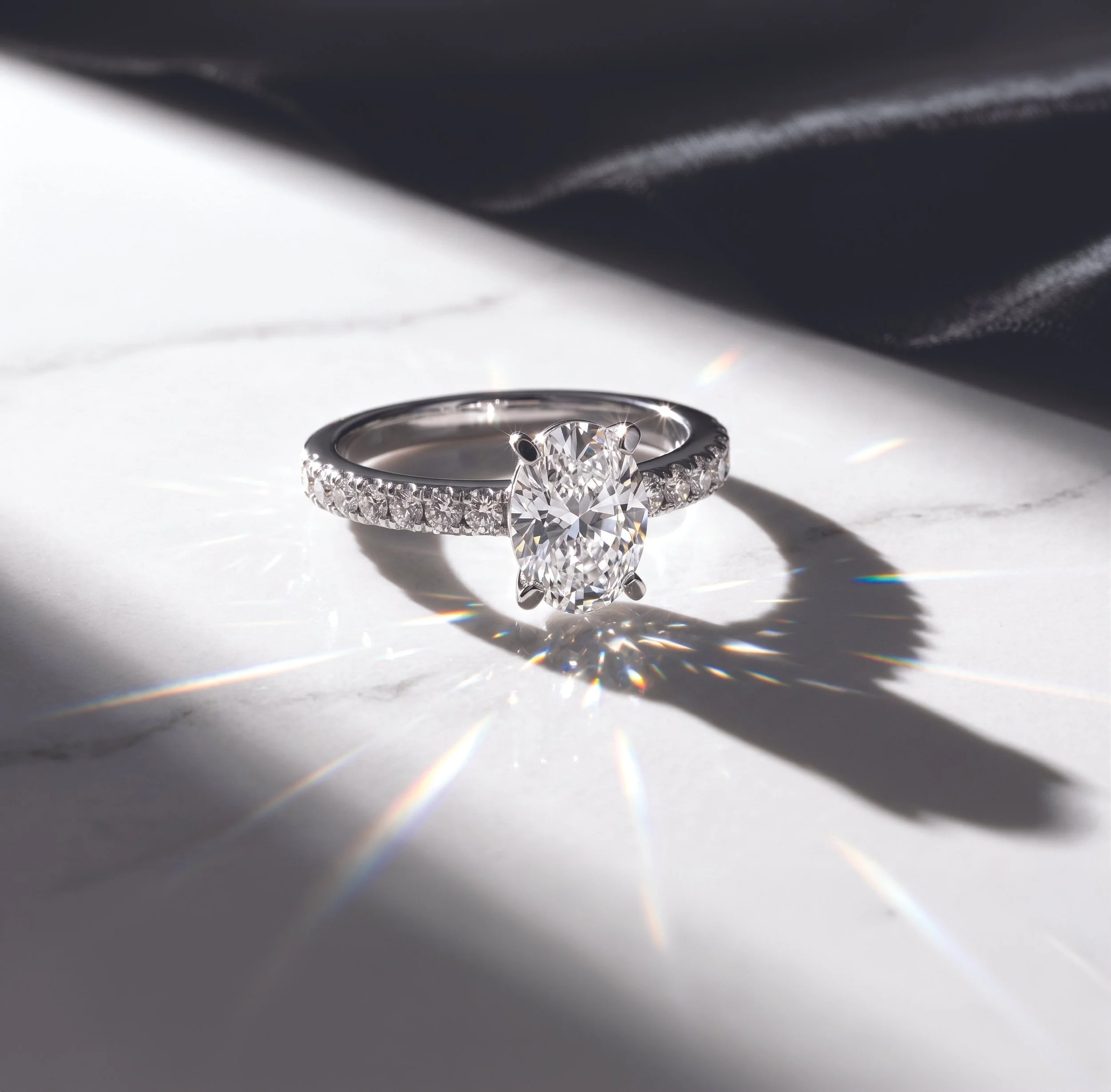 Ein Ring mit einem großen Diamanten in einem Lichtreflex. Der Ring liegt auf einer weißen Oberfläche, die von einem Schatten umgeben ist, während Lichtstrahlen den Diamanten zum Funkeln bringen.