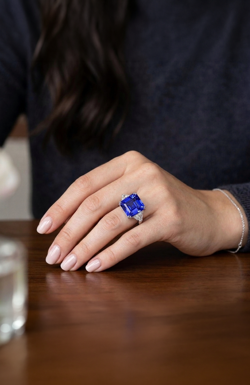 Hand mit einem großen blauen Saphirring auf einem dunklen Pullover, auf einem Holz Tisch, mit einer silbernen Armbanduhr.