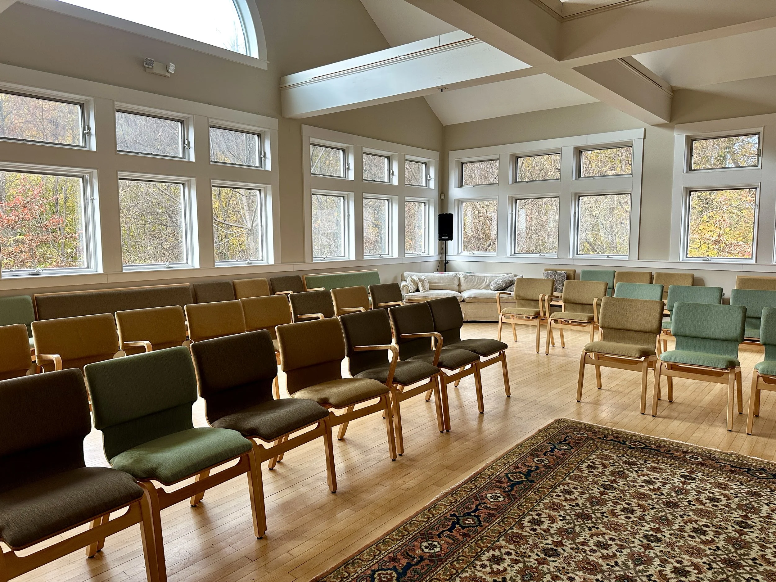 An empty room with rows of green and brown cushioned chairs, large windows showing trees outside, and a patterned rug on a wooden floor.