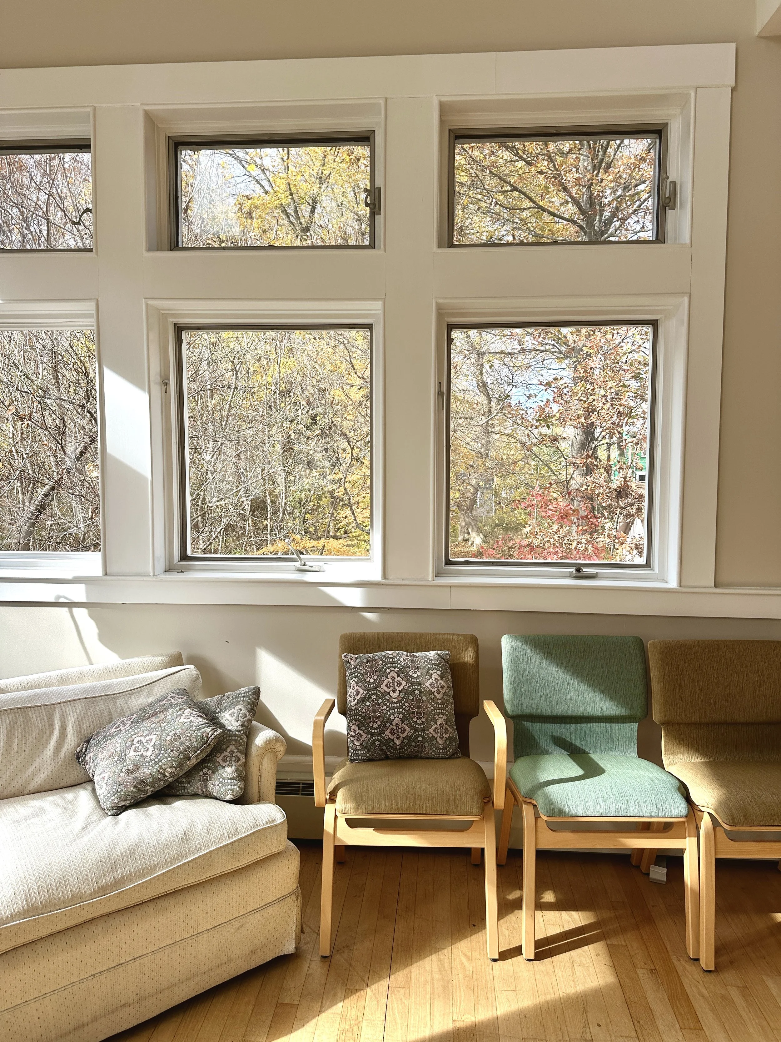 Living room with beige sofa, three chairs with colorful cushions, large windows showing autumn trees outside, hardwood floor, sunlight streaming in.
